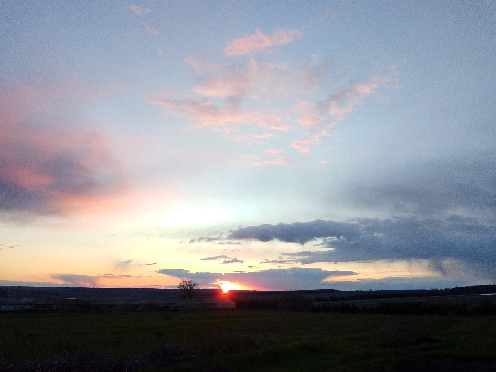 The media file presents a vibrant sunset over a sprawling rural landscape in Khlevnoye, Russia. The sky dominates the upper portion of the image, showcasing a striking gradient of colors. Intense oranges and fiery reds emanate from the setting sun on the horizon, gradually transitioning into soft pinks, light blues, and purples higher up. Thin, wispy clouds in the upper left catch the warm sunlight, appearing pink, while darker, more voluminous clouds are gathered towards the right, with visible rain streaks suggesting localized showers in the distance.
The sun is a brilliant, intense orange-red orb, positioned low on the horizon, partially obscured by a dark tree line or distant terrain. A lone, silhouetted tree stands out to the left of the sun's glow. In the midground, the landscape features a dark, indistinct horizon line, possibly indicative of a flat plain or gentle hills. Several dark, low-lying structures, resembling farm buildings or ruins, are visible in silhouette on the left side of the midground. The foreground consists of dark, grassy ground, with sparse details due to the low light conditions of dusk. No people or visible text are present in the image. The scene conveys a sense of serene natural beauty at the close of day.