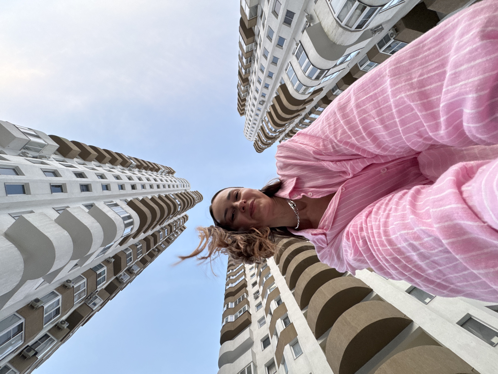 A low-angle, wide-shot captured using a smartphone shows a woman in a pink striped shirt and a silver necklace. She is smiling and looking at the camera. Her long, brown hair is windswept. In the background, the curved balconies and windows of modern apartment buildings are visible against a clear blue sky. The image appears to have been taken in Sochi, Russia, near the sea.