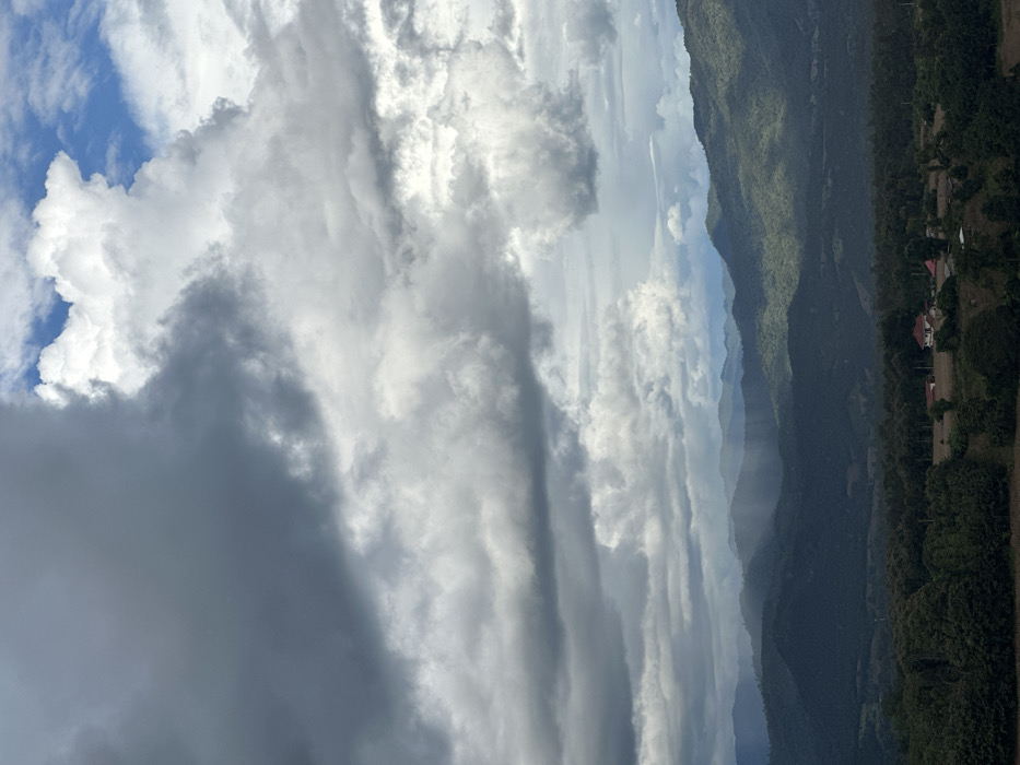 An elevated panoramic view captures a mountainous landscape in Ban Mai Saha Sampan, Thailand. The sky is dominated by a complex cloud system, with dark grey, heavy cloud cover occupying the left portion of the frame, transitioning to brighter white cumulus clouds with visible patches of blue sky towards the upper right. Multiple layers of verdant, tree-covered mountains extend into the background, exhibiting atmospheric perspective. In the mid-distance, distinct shafts of rain are visible descending from the clouds onto a section of the mountains. The immediate foreground, particularly the lower right, features a small cluster of structures, including at least two buildings with reddish-brown tiled roofs, situated amidst cleared or cultivated land and surrounded by dense green trees. No human figures are present.