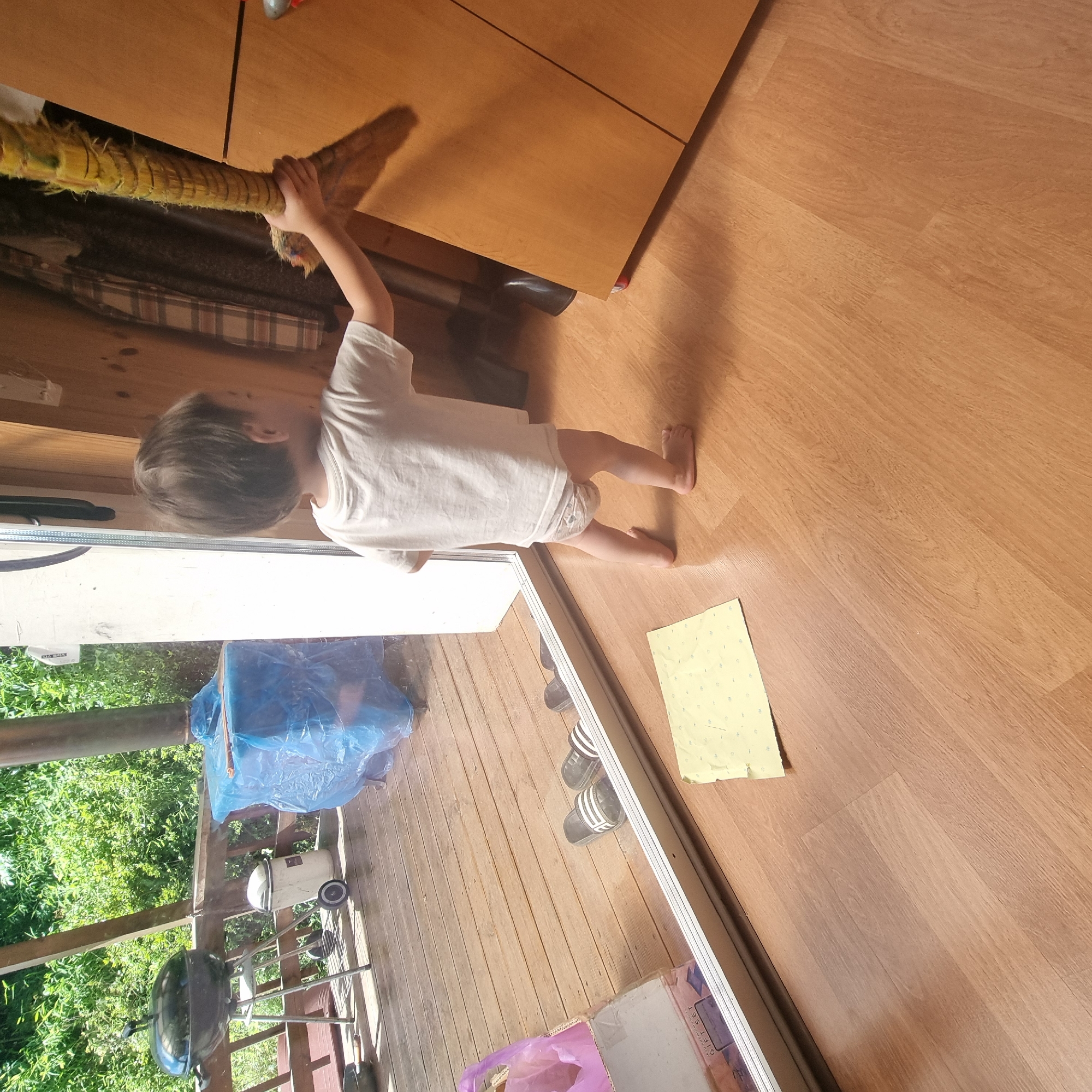 An overhead shot captures a young child, viewed from the rear, standing on a light brown wooden floor indoors. The child, clad in a white t-shirt and a diaper, faces towards a large wooden cabinet on the right side of the frame. The child's right arm is extended upwards, holding a yellow, segmented object that resembles a mace or a large corn cob. Sunlight streams in from a large glass door on the left, illuminating the floor and casting shadows. Through the glass door, a wooden deck with a black barbecue grill, a white trash can, and items covered in blue plastic are visible, suggesting an outdoor space. A small, yellow sheet of paper with blue markings lies on the floor in the foreground. The overall scene appears to be a home environment, possibly a porch or an indoor area connected to an outdoor space.