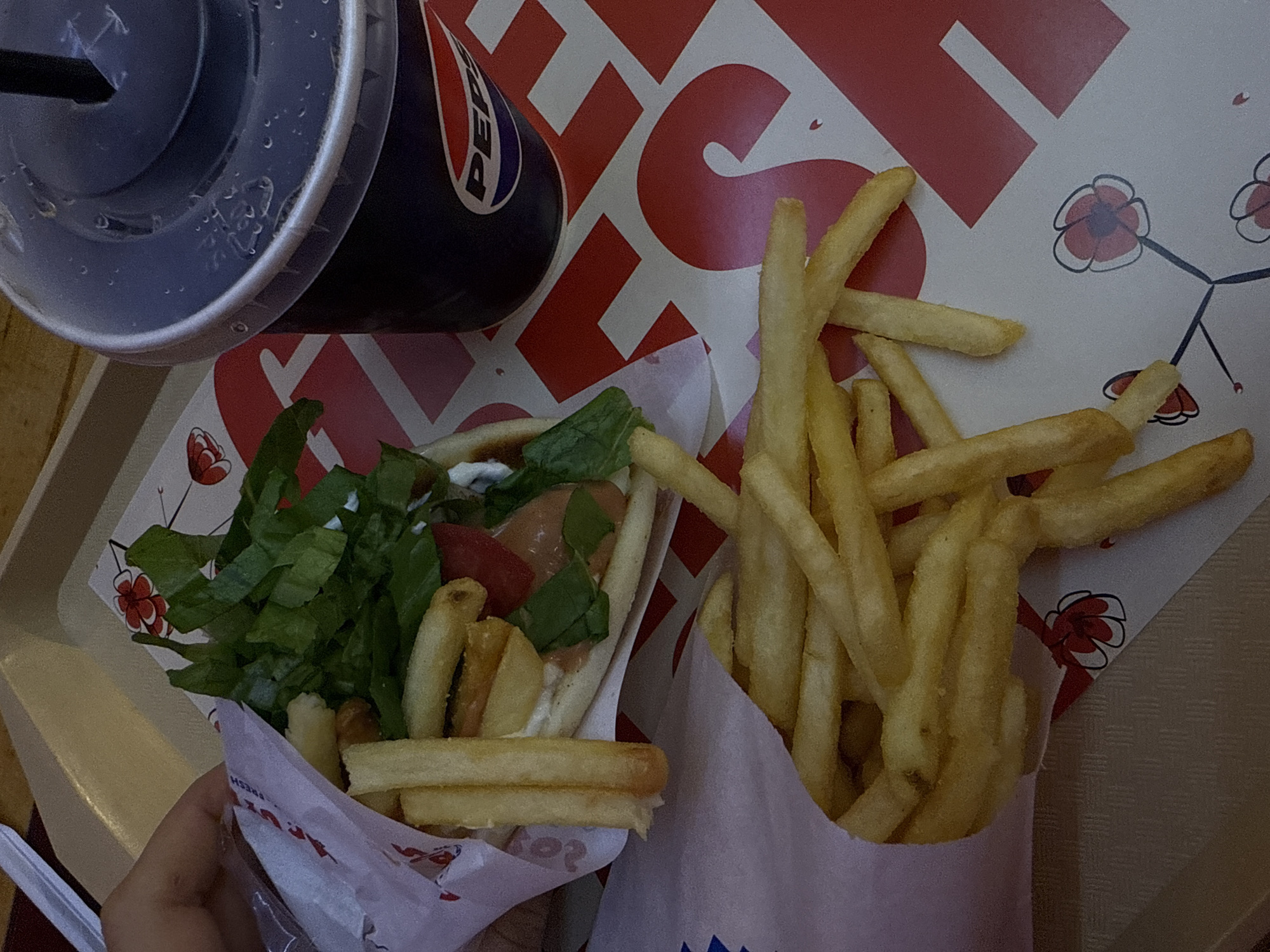 The image depicts a meal, likely consumed at a fast-food restaurant in Yerevan, Armenia. It includes a wrapped gyro filled with fries, lettuce, tomatoes, and what appears to be a creamy sauce. Next to the gyro, there is a separate container filled with more fries. A partially visible Pepsi beverage with a straw is also present. The food and drink are placed on a serving tray. The wrapper of the gyro indicates it could be from a restaurant called "Art Lunch Yerevan". The tray itself rests on a surface with a textured, slightly patterned surface. The overall setting appears to be indoors. A hand can be seen holding the gyro.