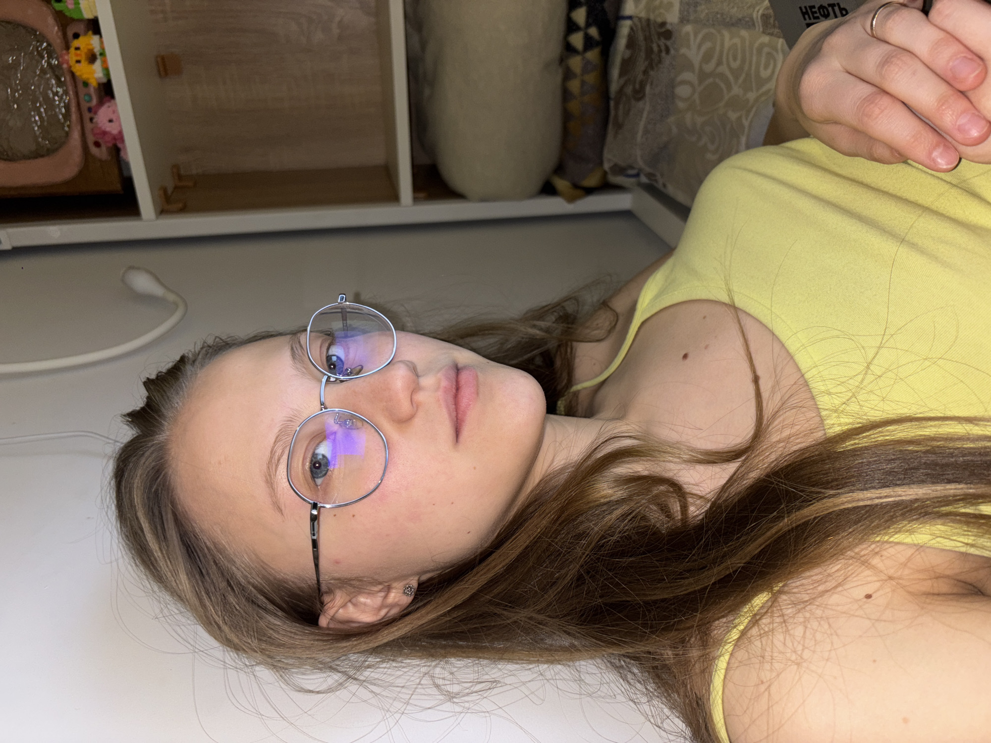 A young, light-skinned individual with long, brown hair and round-framed glasses lies on a white surface. The individual wears a yellow top. A hand with a wedding ring is visible. Behind the individual are light-colored shelves and a patterned cushion. A flexible lamp is visible to the left. The individual appears to be located indoors in Perm, Russia, given the "NEФTb" inscription on the cushion in Russian.
