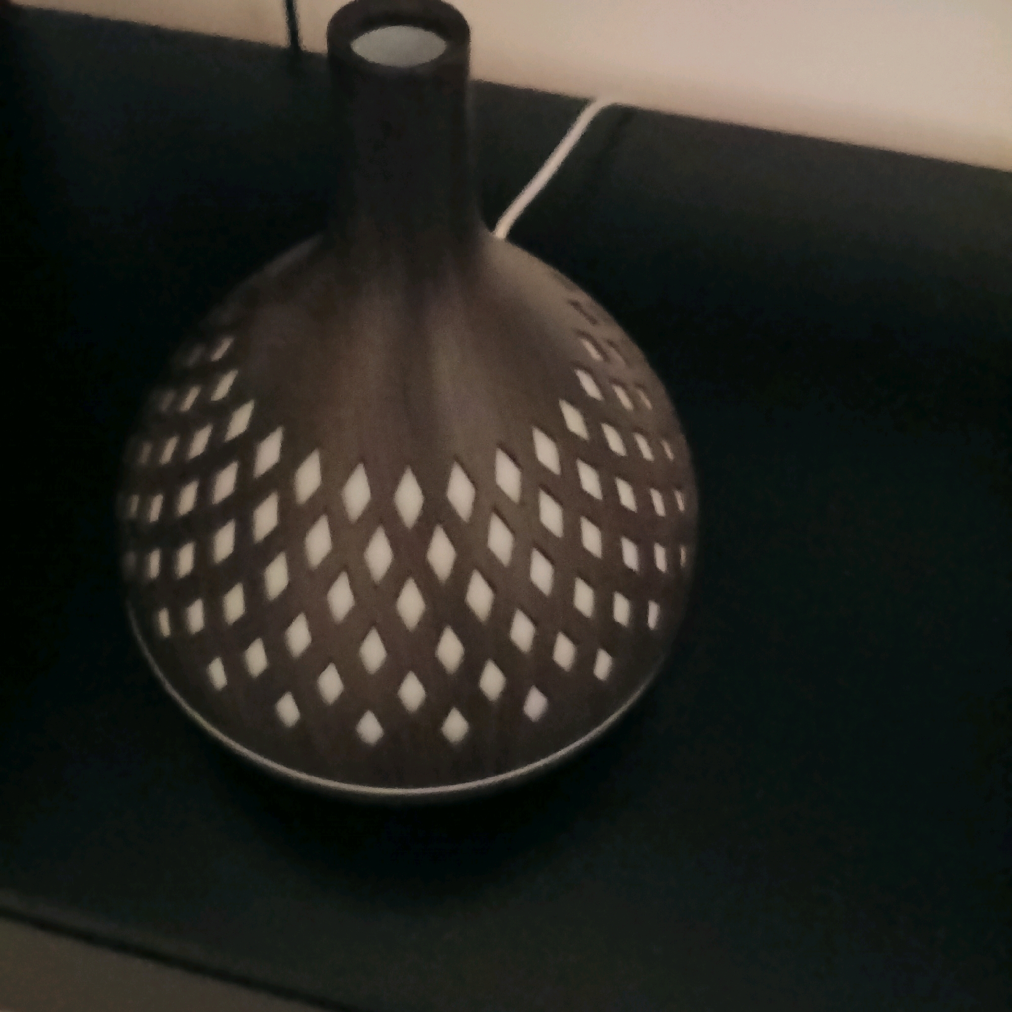 The image shows a close-up, angled view of a dark, round object with a lattice-like pattern of diamond-shaped openings. Through these openings, a white or light-colored interior is visible. The object has a smooth, curved surface and a cylindrical opening at the top. A thin white cord extends from the top of the object and disappears to the right. The object is placed on a dark, matte surface that appears to be a tabletop or shelf. The background is out of focus and consists of dark and light areas, suggesting an indoor environment. There are no people or animals visible in the image. The scene does not contain any elements that would allow for the determination of its location within the city of Bengaluru, India.