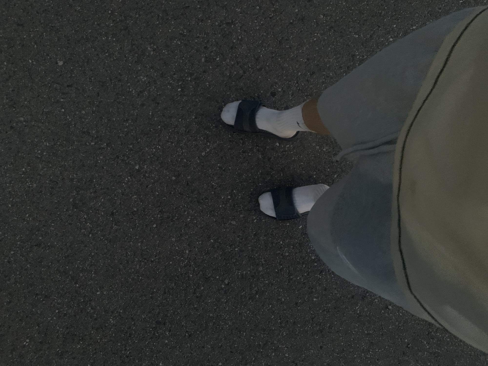 This is a first-person perspective, medium-shot image of a person standing on a dark asphalt surface. The person is wearing light-colored shorts, white socks, and black slide sandals. The asphalt is a rough, textured surface with small, scattered light-colored particles. The lighting suggests it is either early morning or late evening. The scene is devoid of any identifying landmarks or contextual information that would place it specifically in Minsk, Belarus. The focus is solely on the person's feet and lower legs, and the ground beneath them.