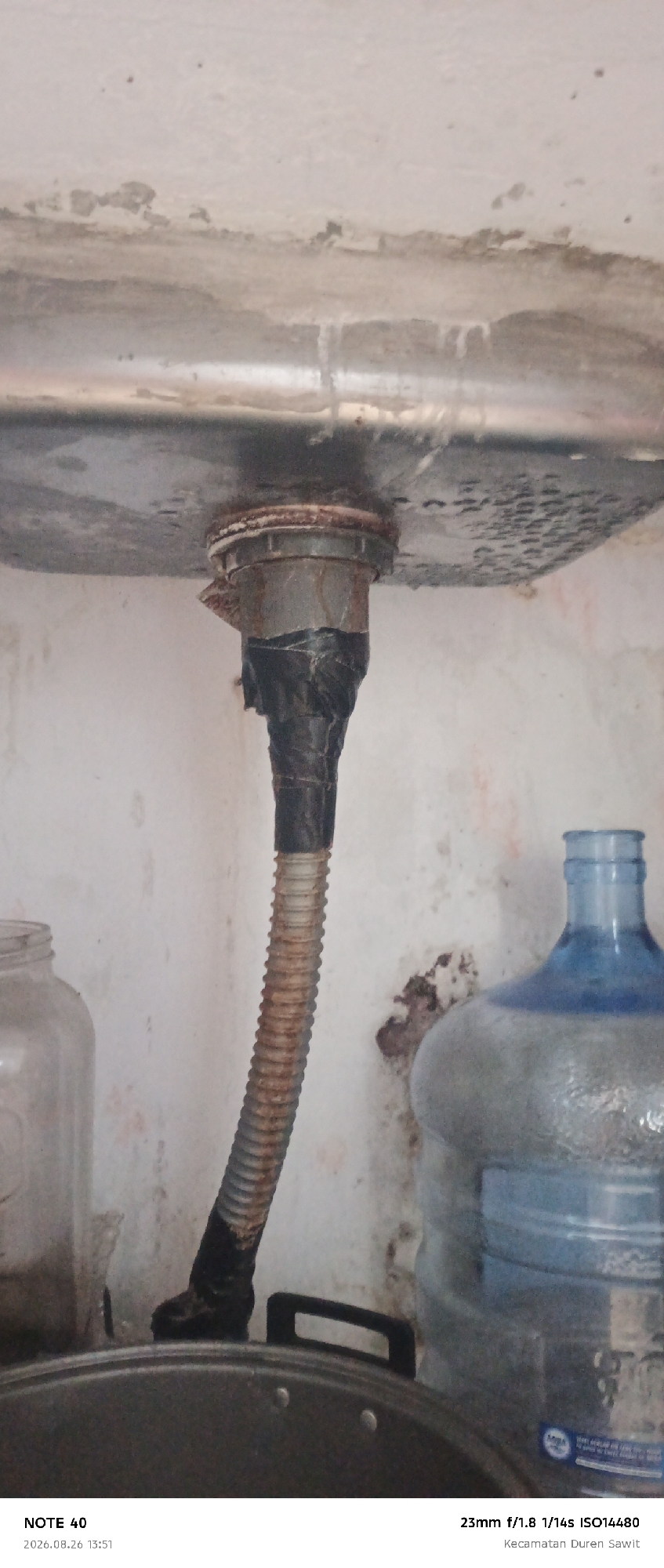 The image shows a close-up of a sink drain with a flexible hose attached. The hose appears to be old and rusted, and it is secured to the drainpipe with black tape. There are also water stains and discoloration on the sink and the surrounding wall. In the background, there is a large plastic water jug. The text at the bottom of the image indicates that the photo was taken on August 26, 2026, at 13:51, and the location is Kecamatan Duren Sawit, East Jakarta, Indonesia. The overall impression is of a somewhat neglected and possibly leaky sink area.