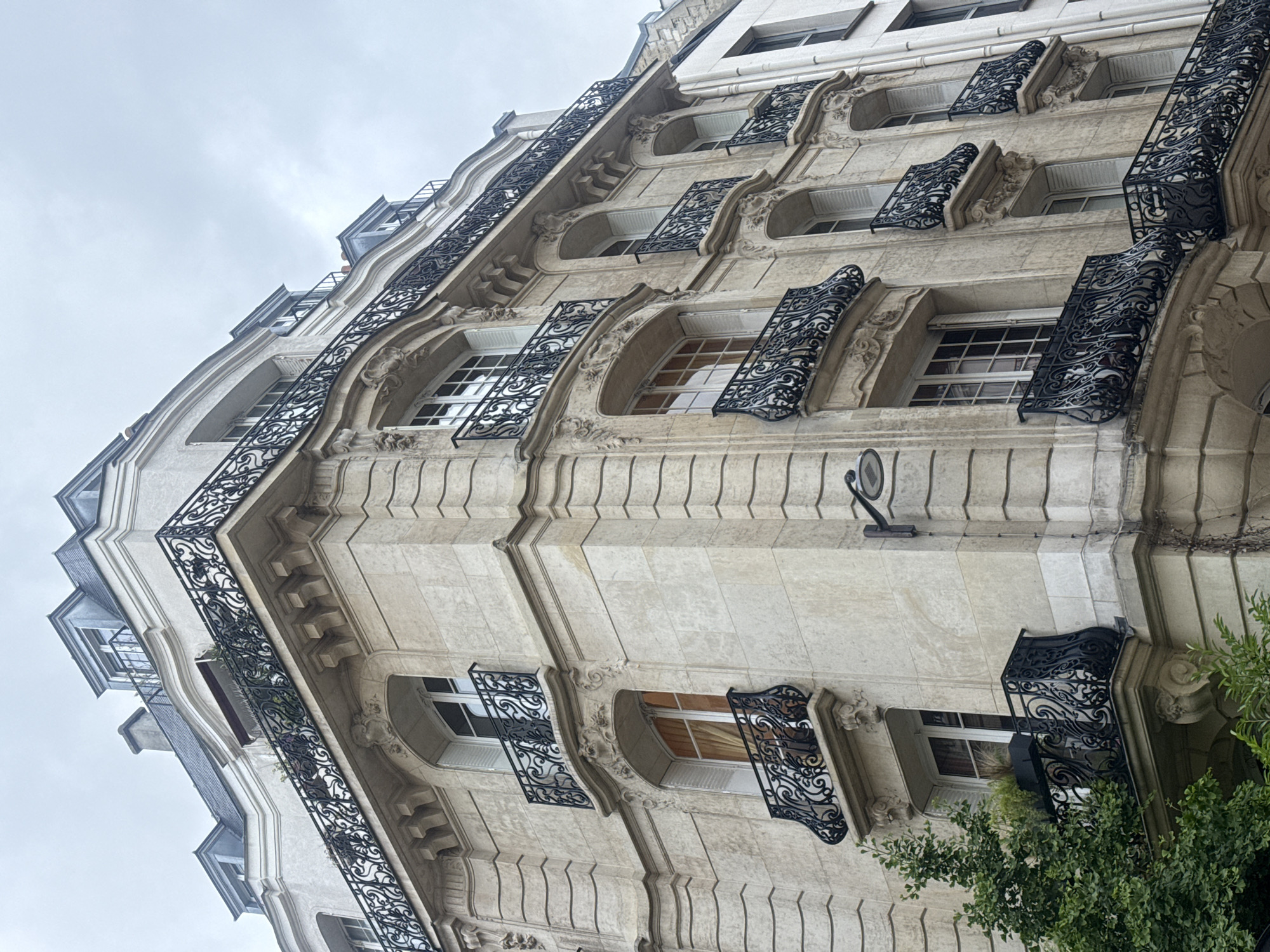 The image shows a low-angle view of a Parisian building facade. The building is made of light-colored stone with ornate detailing around the arched windows. Black wrought-iron balconies are attached to each window. The sky is overcast. The architecture is typical of buildings found in Paris. Green foliage is visible on the right side of the building.