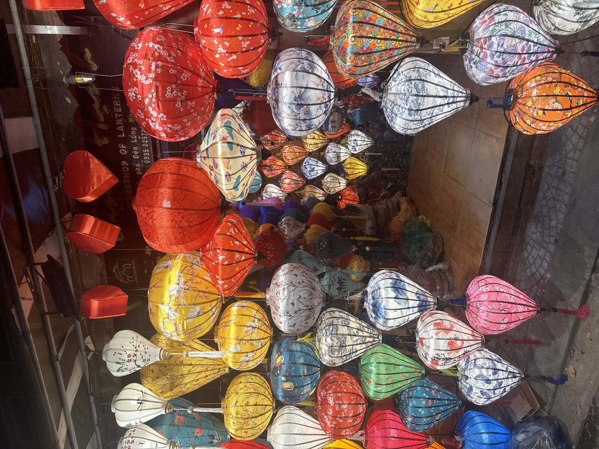 The image depicts a shop displaying colorful lanterns in what is possibly a city in Vietnam. The lanterns are of various shapes, sizes, and colors, predominantly red, yellow, white, and blue, many featuring floral or bird patterns. Some lanterns are hung close to the shop’s facade, casting a warm glow, while others are arranged deeper inside the shop. A sign labeled "WORKSHOP OF LANTERNS" with a phone number is partially visible in the background. The ground or street visible at the bottom of the frame suggests the shop is street-side. The overall scene gives a sense of a vibrant and traditional craft business.