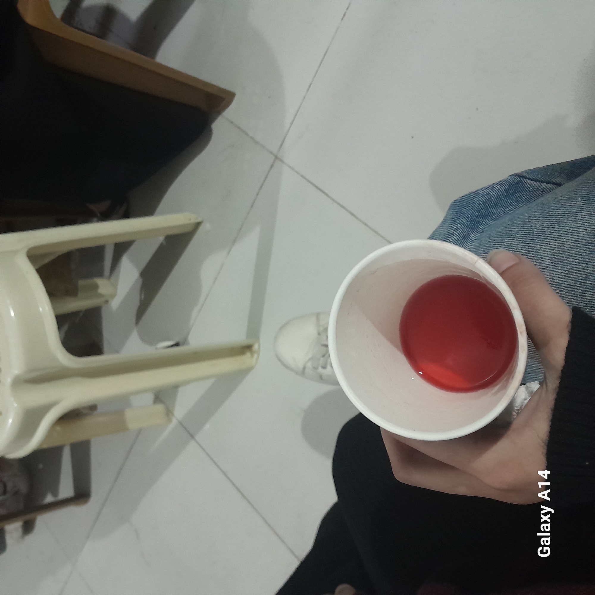 The image shows an overhead perspective of a person sitting in a room. The person is holding a white paper cup filled with a red liquid. They are wearing blue jeans and a black long-sleeved shirt. A white sneaker is partially visible near the cup. Next to the person is a white plastic high chair and a brown and black chair. The floor is tiled. There is no information available in the image about the location of the image, including the city of Khan Yunis, Palestinian Territory.
