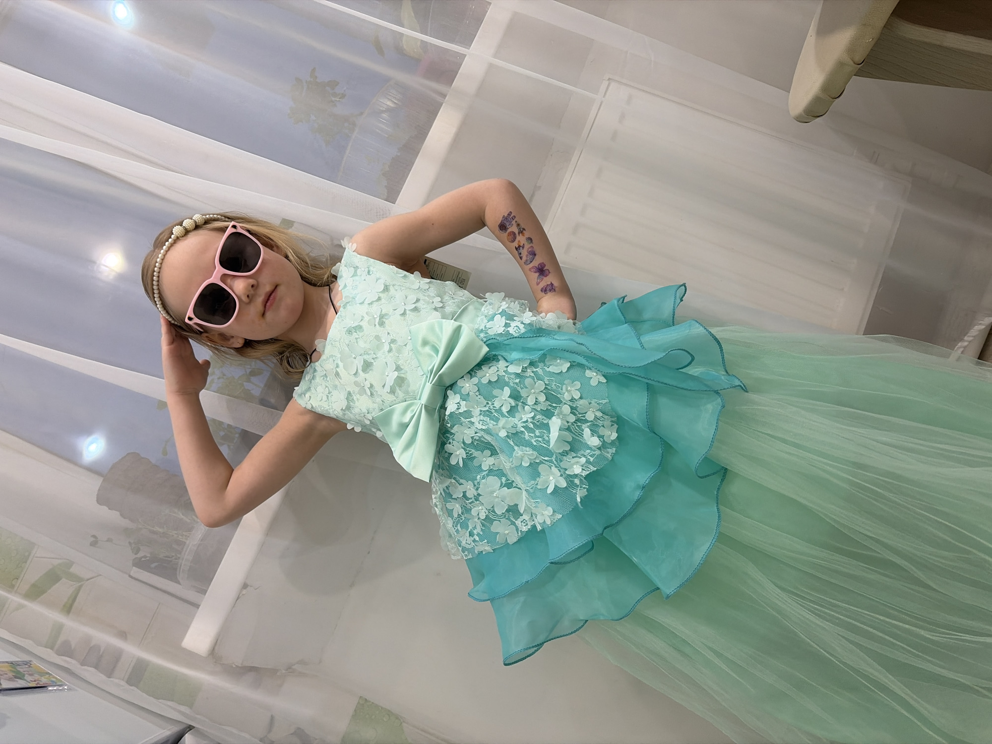 A young girl is captured indoors, posing confidently in a slightly tilted, medium shot. She wears a formal, two-toned dress that transitions from a light mint green bodice to a flowing, tiered skirt. Her blonde hair is adorned with a pearl-beaded headband, and she sports pink square-framed sunglasses that obscure her eyes. Her right hand rests near her head, subtly adjusting the sunglasses, while her left hand is placed on her hip. Visible on her right forearm are several colorful temporary tattoos, including butterflies and a snail.
The dress features a light mint green bodice with an intricate white lace overlay embellished with small white floral appliques. A large, light mint green bow is prominently tied at the waist. The skirt flows down from the bow with tiered ruffles in a slightly darker turquoise or teal color, transitioning into a full, sheer, light mint green tulle skirt that extends to the floor.
The background primarily consists of sheer white or off-white curtains, likely covering a window, which diffuse bright light and create soft reflections. Fragments of light-colored furniture, possibly a chair or table edge, are visible in the upper right and lower left corners of the frame. On a white surface in the bottom left, a small item resembling a book or magazine shows part of a word, possibly "Drea" or "Dream." A small tag on her dress shows the number "34."
The scene takes place indoors, likely a home or a clothing store, given the elaborate dress and clean setting. The lighting suggests a bright environment, though the specific time of day is not discernible. The girl appears to be confidently posing, exuding a playful and stylish demeanor. The location context provided is Noviy, Russia, though no specific visual cues in the image confirm this.