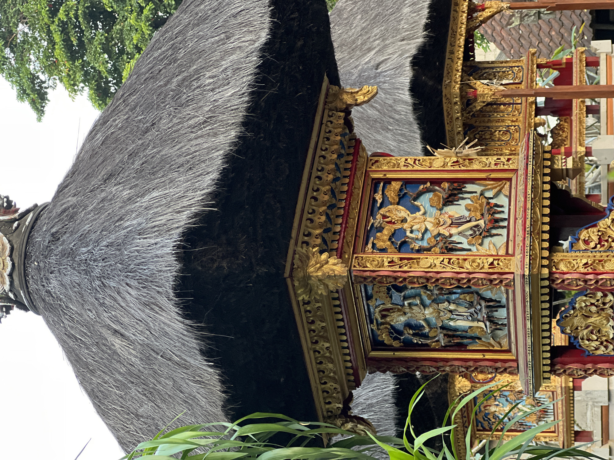 A close-up, eye-level shot captures the intricate details of traditional Balinese architecture in Ubud District, Indonesia. The foreground features a tiered structure with a thatched roof, its golden ornamentation and carved panels highly detailed. These panels depict figures in traditional attire, set against vibrant blue backdrops, possibly representing deities or mythological scenes. The roof, made of dry grass, is thatched in overlapping layers, creating a textured surface. In the background, another similar structure with a thatched roof is partially visible, along with glimpses of lush green foliage and terracotta-tiled rooftops, suggesting a temple complex or a traditional Balinese compound. The lighting suggests it is daytime.