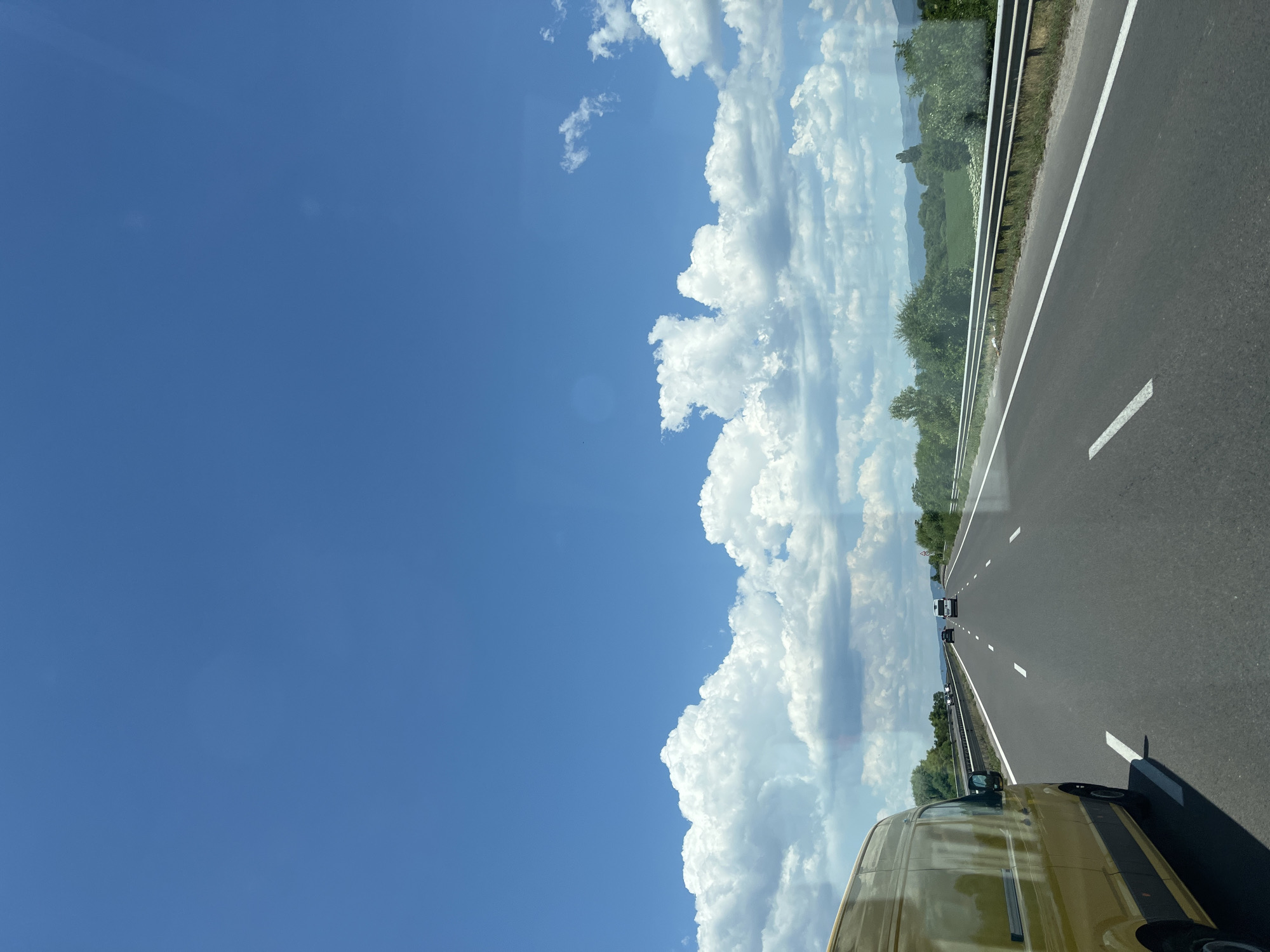 The image depicts a view from inside a vehicle traveling on a highway. A yellow van is visible in the foreground on the left side of the frame. The highway stretches into the distance, with white lane markings. Other vehicles are present on the road ahead. The sky is a bright blue with scattered white clouds. The landscape on either side of the highway consists of green fields and trees, with rolling hills or mountains in the background. The scene appears to be set in a rural or suburban area during the daytime.