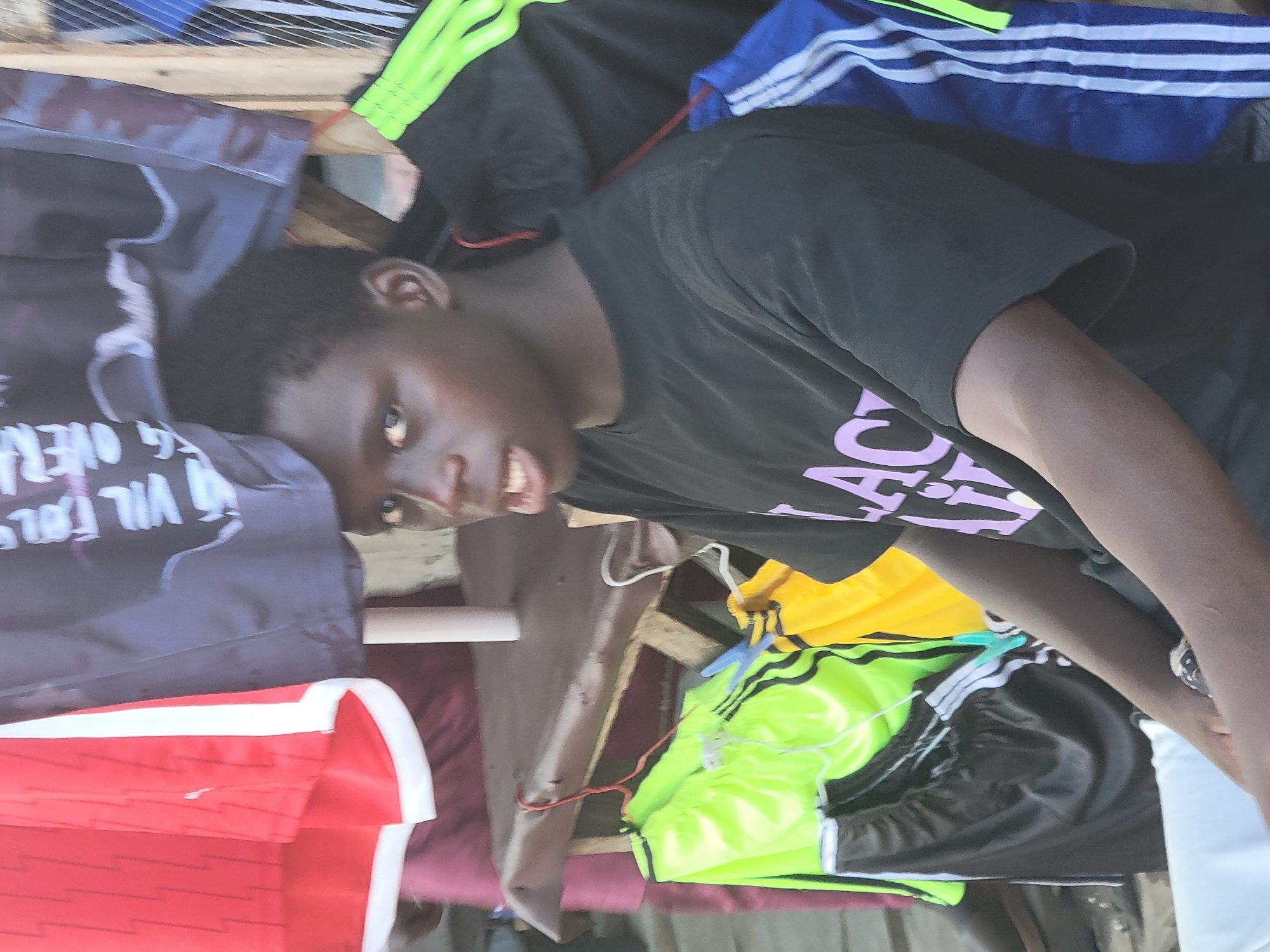This image, taken in Madalla, Nigeria, depicts a young Black person amidst what appears to be an outdoor market stall. The photograph is rotated 90 degrees clockwise.
The central subject is a young individual with short, dark curly hair and dark skin. They are looking directly at the camera with a slight smirk or smile, revealing a hint of their teeth. They are wearing a black short-sleeved t-shirt with prominent light purple or pink text. The visible text clearly spells "BLACK LIVES" and the partial word below it appears to be "MATTER". A light-colored band or watch is visible on their right wrist.
The person is surrounded by an assortment of clothing items, indicating a market setting. To their right, several sportswear items are displayed, including dark blue track pants or shorts with white stripes (reminiscent of Adidas) and bright neon green shorts with black stripes. A yellow garment and a black one are also present. To their left, a dark grey or black fabric item, possibly another t-shirt, features white, upside-down text that reads "VIL FOL..." and "OVER". Further to the left and below, a folded red garment, possibly a jersey, with white trim or stripes is visible.
The background reveals a simple market setup with wooden planks that form part of a stall or shelving, and some grey mesh material at the top. White plastic hangers are used for display, and a thin red string or wire is noticeable near the center. The lighting suggests it is daytime. The scene conveys a casual, informal market atmosphere.