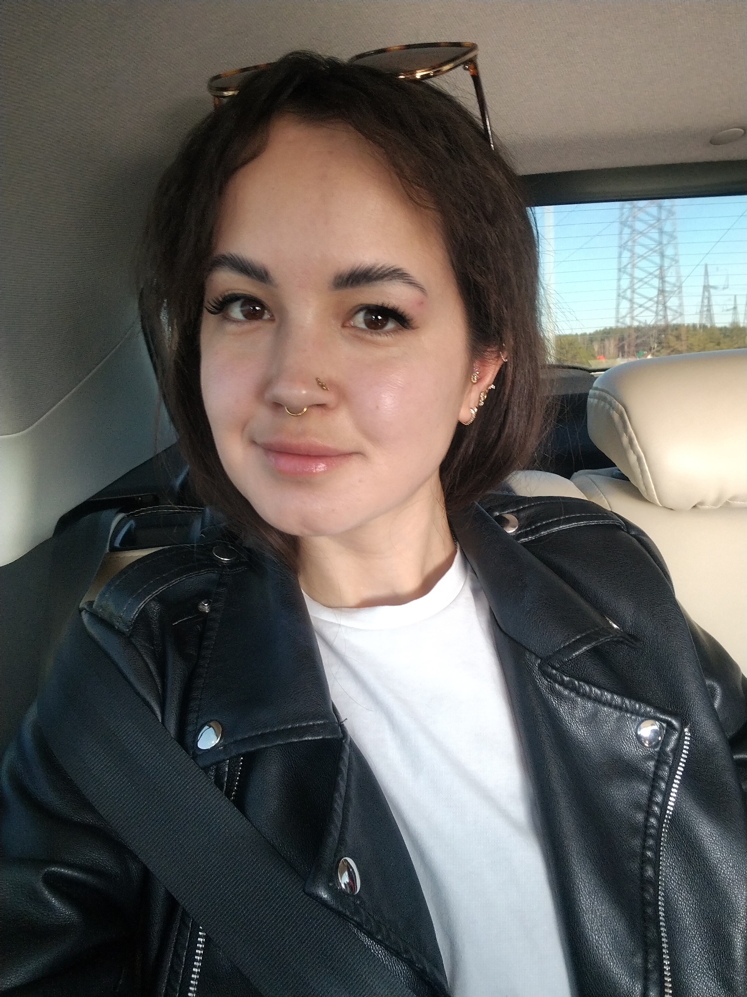 A selfie depicts a woman in the backseat of a car. She is wearing a black leather jacket over a white t-shirt and has a seatbelt across her torso. Sunglasses are perched on top of her head. She has dark hair, brown eyes, a nose ring, and multiple earrings in her left ear. Outside the car window, transmission towers can be seen on the horizon against a blue sky with some visible trees. The location is Noviy Urengoy, Russia.