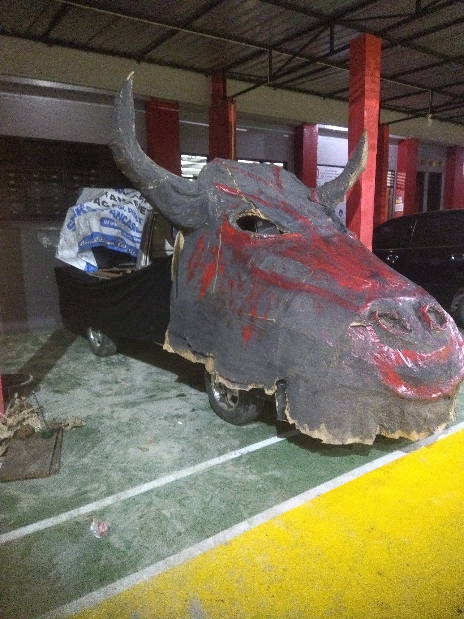 The image shows a large, homemade bull costume covering the rear of a three-wheeled vehicle parked indoors, possibly in a garage or covered area. The costume is primarily gray with red paint markings to simulate a bull's face and blood. It has two horns and eye openings. The vehicle is partially covered with black fabric and a white tarp with text that appears to be in Indonesian. The ground is concrete with green and yellow painted lines. Red columns are visible in the background, along with a black car. The setting appears to be an industrial or service area. No people are visible in the image. The specific location in Giritirto, Indonesia, is not discernible from the visual information alone.
