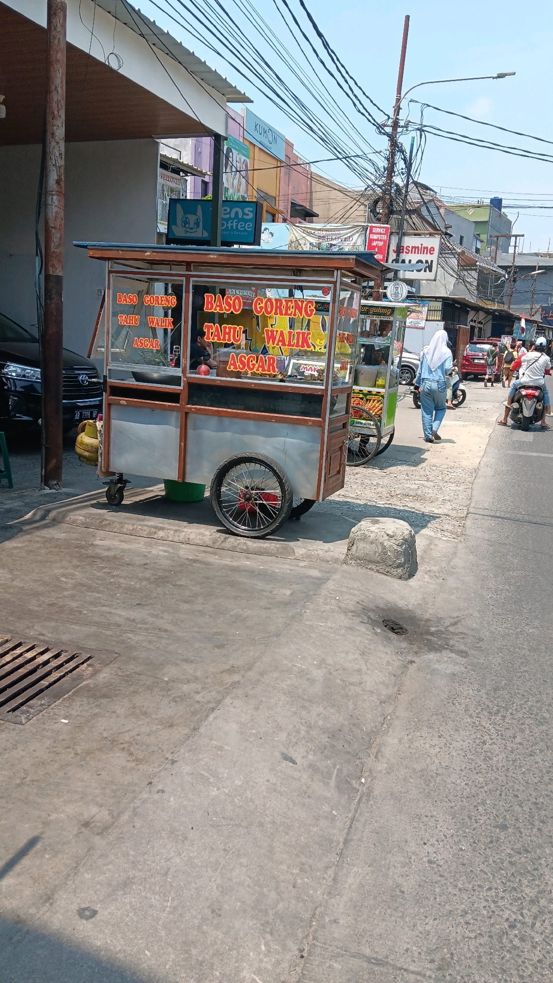 The image captures a street scene in Depok, Indonesia, focusing on a mobile food cart specializing in "Baso Goreng" (fried meatballs) and "Tahu Walik" (inside-out tofu). The cart, made of wood and metal, is equipped with glass display cases showing the food and signage advertising its offerings in Indonesian. A yellow gas tank is attached to the side of the cart. Behind the cart, a black car with the license plate "AB 1306 WP" is parked.
In the background, a variety of buildings line the street, displaying commercial signage. Notable signs include "KUMON," "ENS COFFEE," "PRELOVED," and "JASMINE SALON." The sky is clear and bright, suggesting a sunny day. Power lines crisscross overhead, a common sight in urban Indonesian environments.
Several individuals are visible in the scene. A person wearing a light blue hijab and pants walks away from the camera towards the food cart. Further down the street, two people are riding a motorcycle, and another person is seen walking. The foreground shows a paved sidewalk and road, with a metal grate for drainage. The overall atmosphere suggests a bustling, everyday street scene in an Indonesian city.