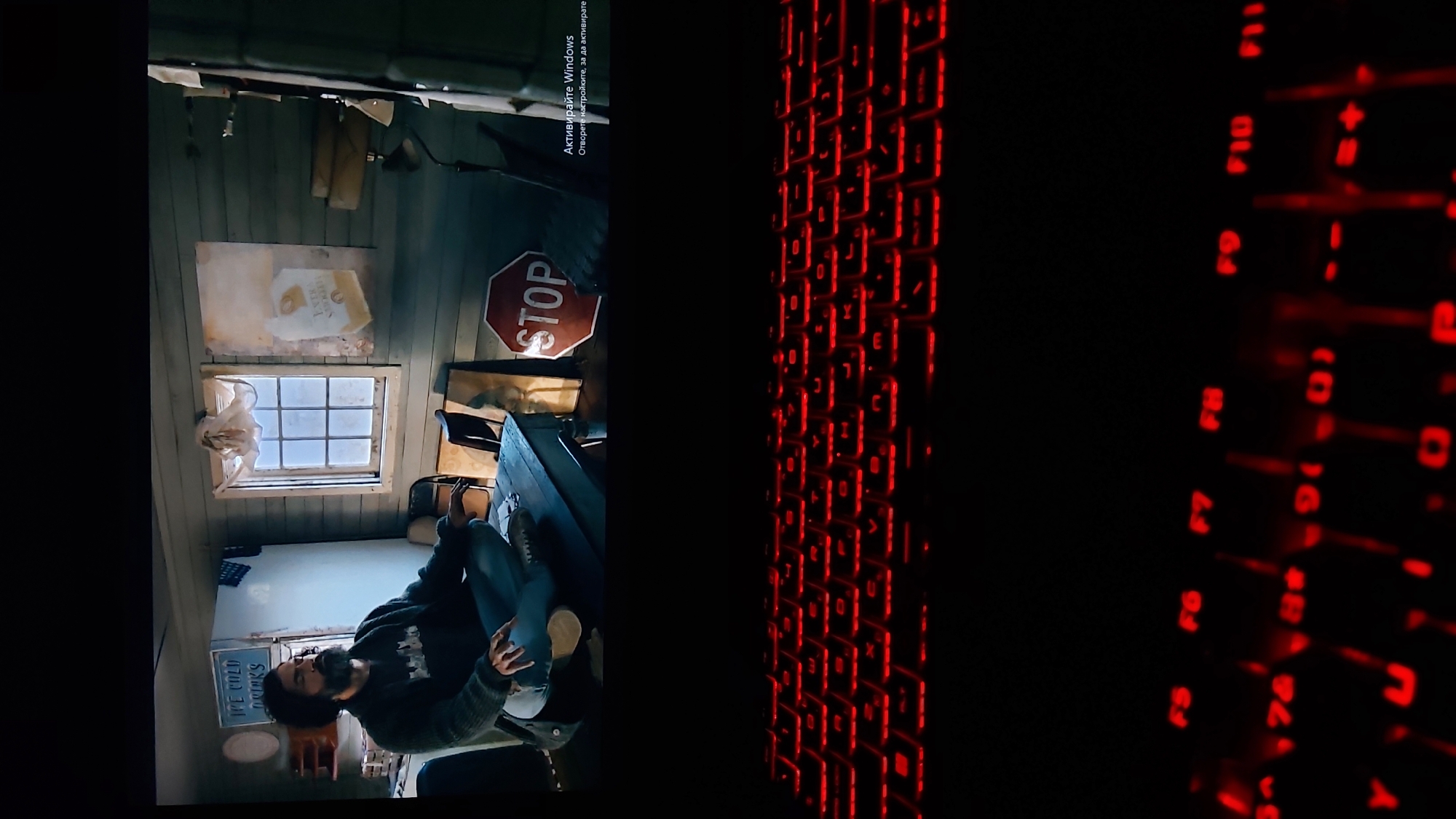 The image shows a scene from a television or computer screen, with a red-lit keyboard filling the right half of the frame. On the left, the screen displays a man sitting in what appears to be a rundown or rustic interior space. There is a window with a white curtain, and a stop sign is hung on the wall. A sign that says "Ice Cold Drinks" is also visible. In the top right corner of the screen, there is a notification saying "Activate Windows" in Bulgarian.
