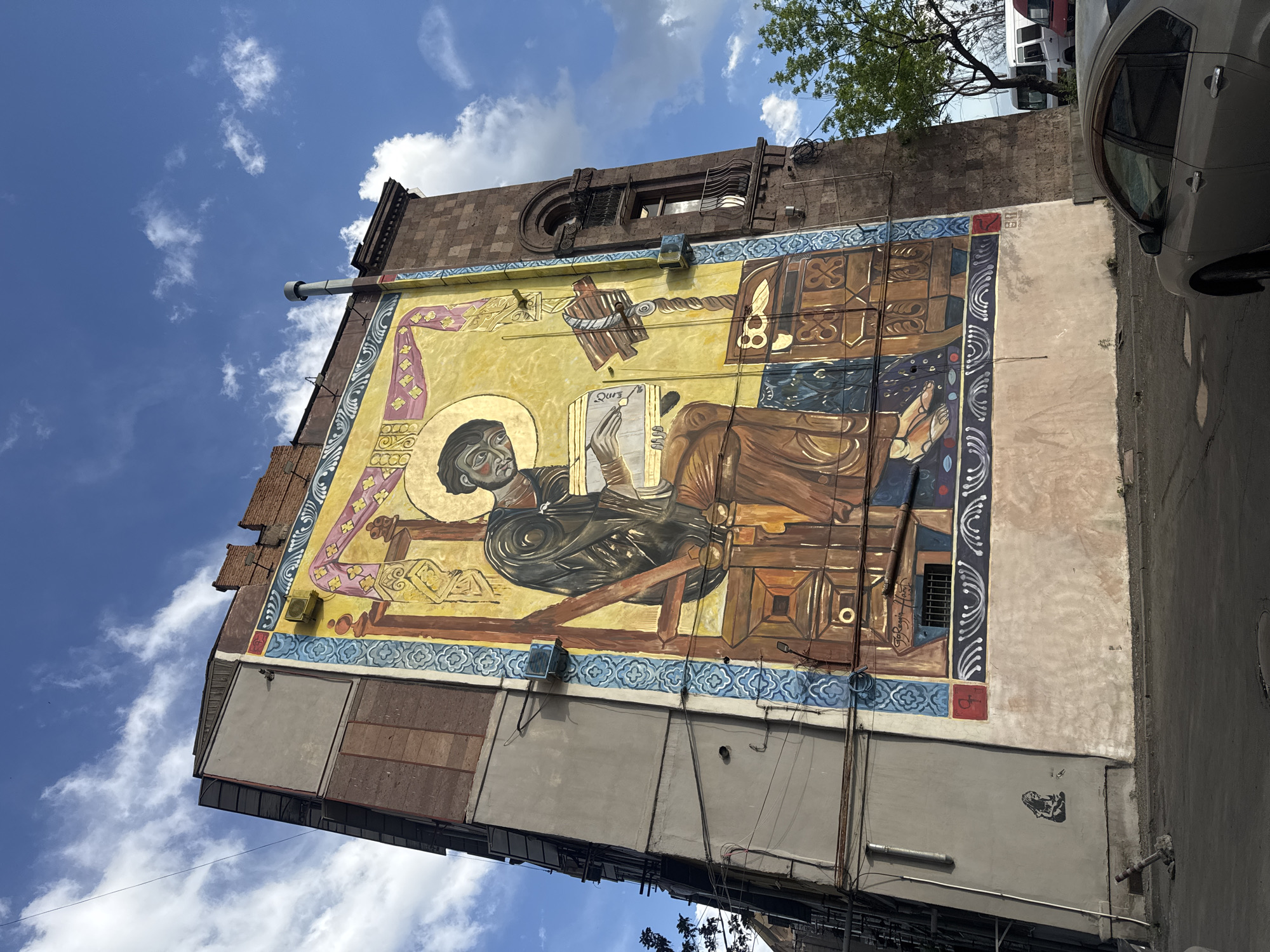 The image captures a mural on the side of a building in Yerevan, Armenia. The mural depicts a religious figure, possibly a saint or scholar, writing in a book. The figure has a halo and sits on a chair with a desk or table in front of them. The style of the mural appears to be inspired by traditional Armenian iconography.
The mural covers a significant portion of the building's exterior wall, extending from a gray and brown lower section to the upper part near the roof. Above the mural, the building's architecture includes details like a small window and decorative elements. The sky is visible in the background, with scattered clouds. Part of a car is visible on the right side of the image, suggesting the building is situated on a street. Vegetation (a tree) also features in the shot.