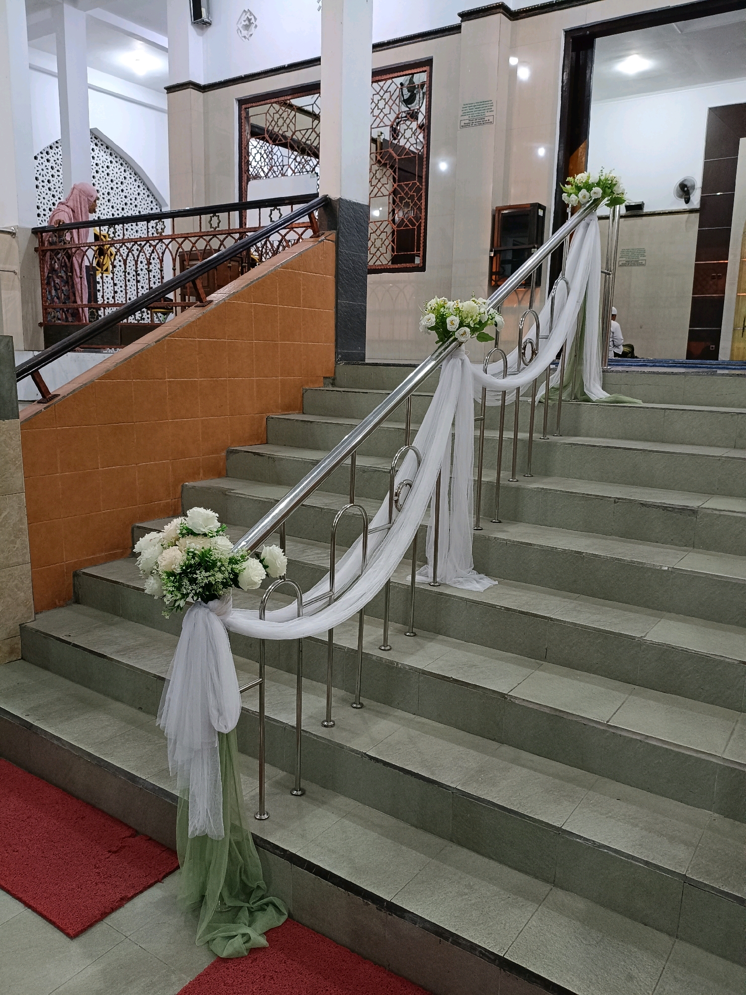 The image depicts the interior staircase of a building, likely a mosque or a similar public venue, in Maguwoharjo, Indonesia. The staircase is decorated with white and green draped fabric and bouquets of white flowers along the metal railing. The steps are made of grey tiles, and the wall to the left is tiled with orange. A person wearing a pink hijab is visible on an upper level, looking over the railing. In the background, through an open doorway, another person in white attire is seen sitting on a blue carpet. The environment appears to be indoors with artificial lighting.