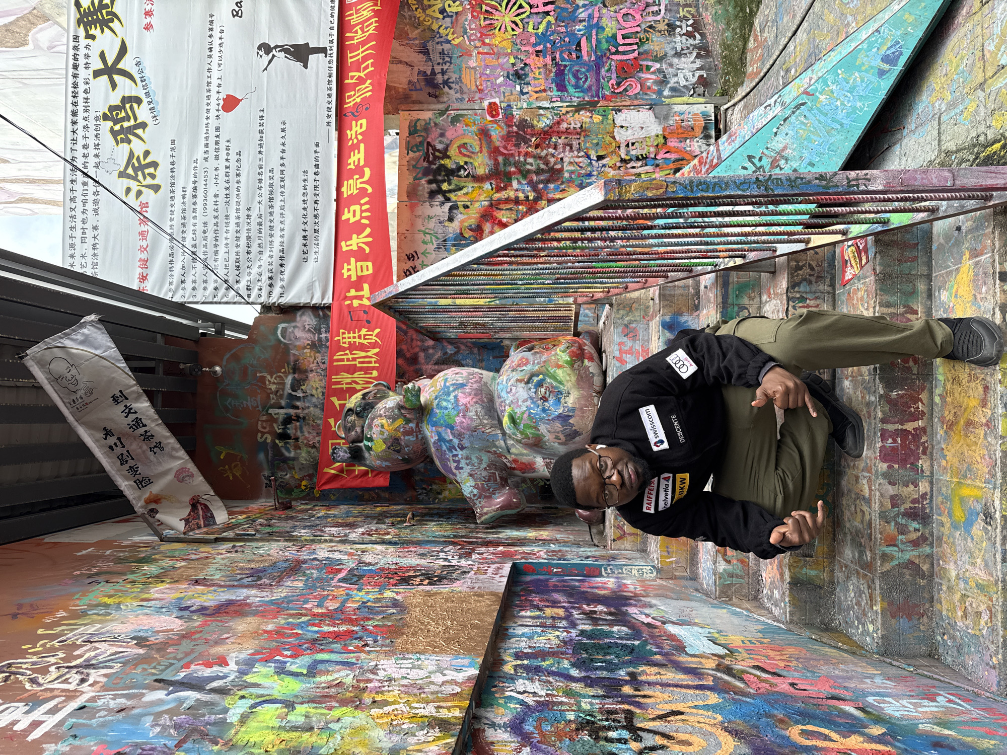 A Black male subject is depicted crouching on a multi-colored, graffiti-covered concrete staircase in an urban alleyway within Jiulongpo District, China. The subject, positioned centrally, wears a black jacket with various corporate logos including "RAIFFEISEN," "helvetia," "swisscom," "DESCENTE," "BKW," and "Audi," along with olive-green cargo pants and black athletic shoes. He wears glasses and white wireless earbuds, and maintains direct eye contact with the viewer while holding both hands in a "hang loose" gesture.
The environment is characterized by extensive, vibrant graffiti covering all visible surfaces: the walls, staircase steps, and a diagonal turquoise ramp structure on the right. Behind the subject, a stack of three abstract, multi-colored sculptures, appearing to be rounded figures, is visible. Above the sculptures and to the right, a red banner with white Chinese characters is displayed horizontally. Further to the right and higher, a large white banner with black Chinese text and a black silhouette of a figure (possibly a child) is affixed to a wall. A white flag with black Chinese script and illustrations is draped from an upper structure on the left. A metal handrail with individually multi-colored painted bars runs along the right side of the staircase. The scene conveys a highly decorated, artistic public space.