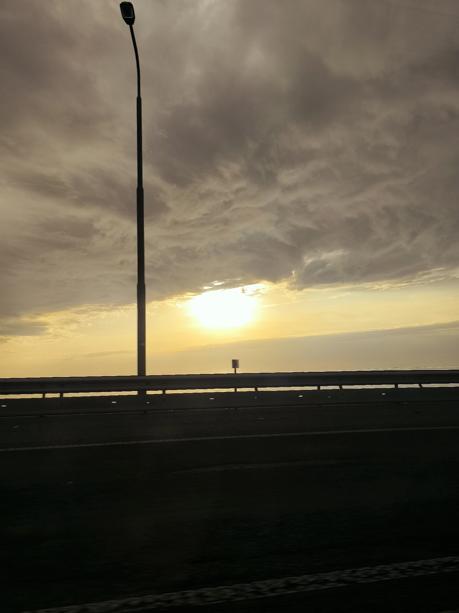 A street lamp stands tall against a dramatic, cloudy sky at sunset. The sun's bright light pierces through a gap in the clouds, casting a warm glow over the horizon. In the foreground, a highway barrier separates the road from the sea, with the dark asphalt of the road visible in silhouette. The scene suggests a location near the coast, possibly a bridge or an elevated road overlooking the water. The overall atmosphere is serene and contemplative, with the natural beauty of the sunset contrasting with the man-made structure of the lamp and barrier. The specific location in Lelystad, Netherlands, could be identified through contextual clues such as unique bridge designs, specific types of street lamps, or distinctive coastal features, which are not clearly discernible from the image alone.