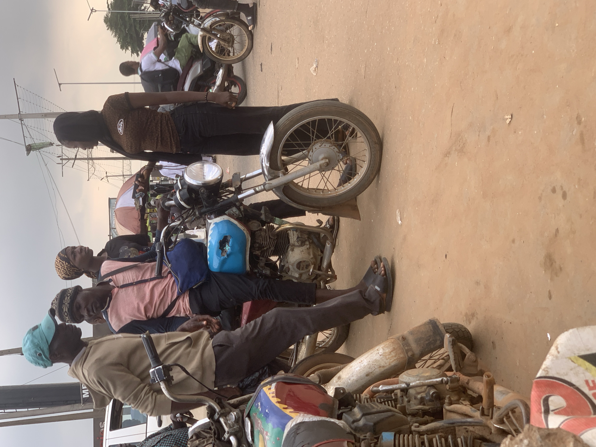 In Shagamu, Nigeria, a group of people are gathered around motorcycles on a dusty, unpaved road. The sky is overcast, and overhead wires crisscross the scene. In the foreground, a man wearing a light brown hoodie and a teal cap is leaning against a motorcycle. Beside him, another man in a pink shirt and a patterned beanie is seated on a motorcycle. A woman with a patterned headscarf stands behind them. Further back, a young woman in a dark, patterned top and black pants stands facing left, holding a phone. Several motorcycles are visible, some decorated with colorful designs. In the background, a white vehicle and other people are present, along with market stalls or umbrellas. The ground is a mix of dirt and sand.