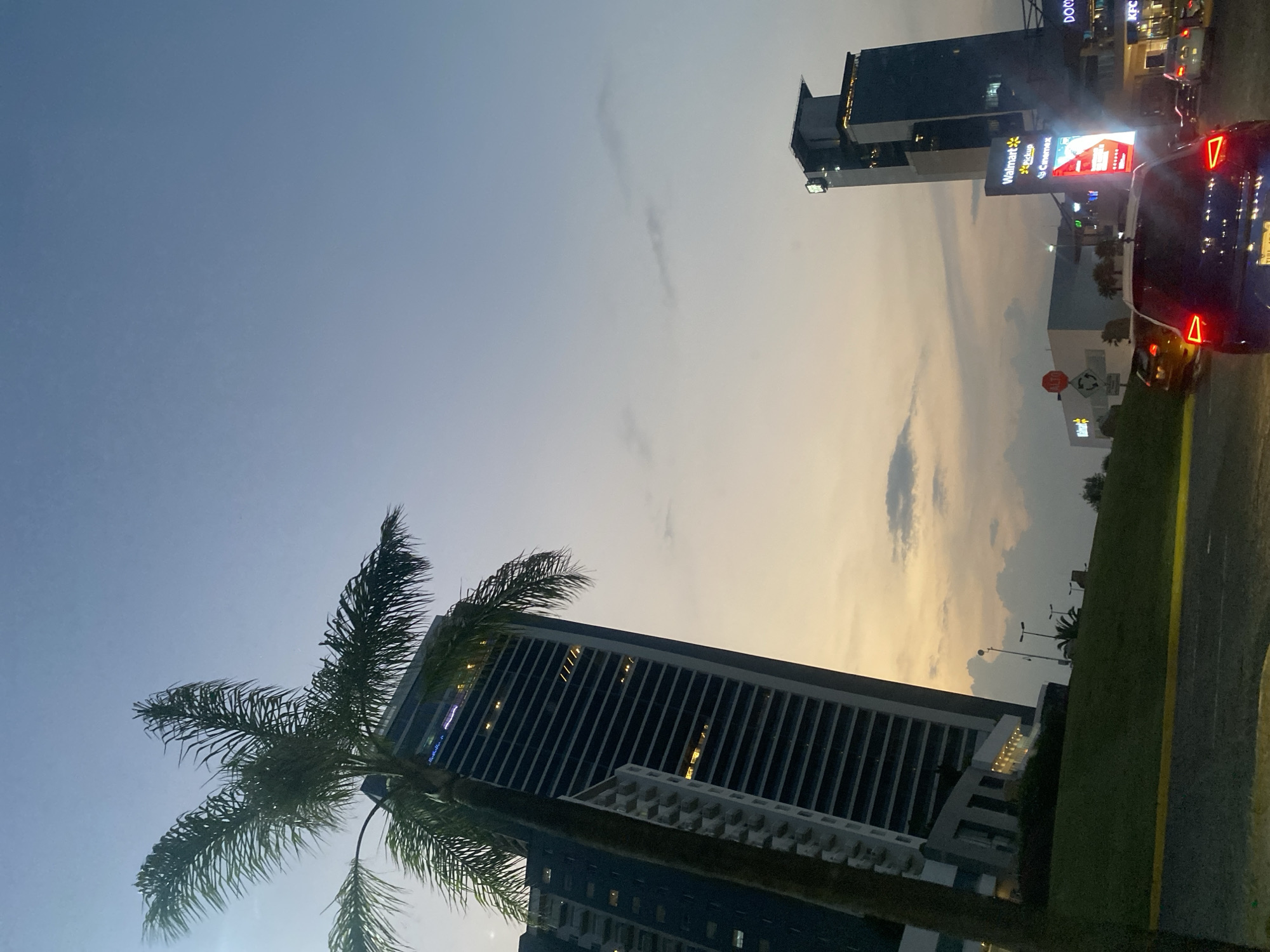 The image shows a cityscape at dusk. A tall, dark-colored skyscraper with vertical lines dominates the bottom center of the frame, partially obscured by a palm tree in the foreground. Above and to the right of this building, another taller building with multiple floors and signs for businesses, including "Walmart Pickup," "Cinemex," "DOGOS," and "KFC," is visible. Cars are lined up on a road running along the right edge of the frame, their red taillights illuminated. A strip of green vegetation separates the road from the buildings. The sky is a mix of pale blue and orange hues, indicating the transition from day to night, with streaks of clouds visible.
