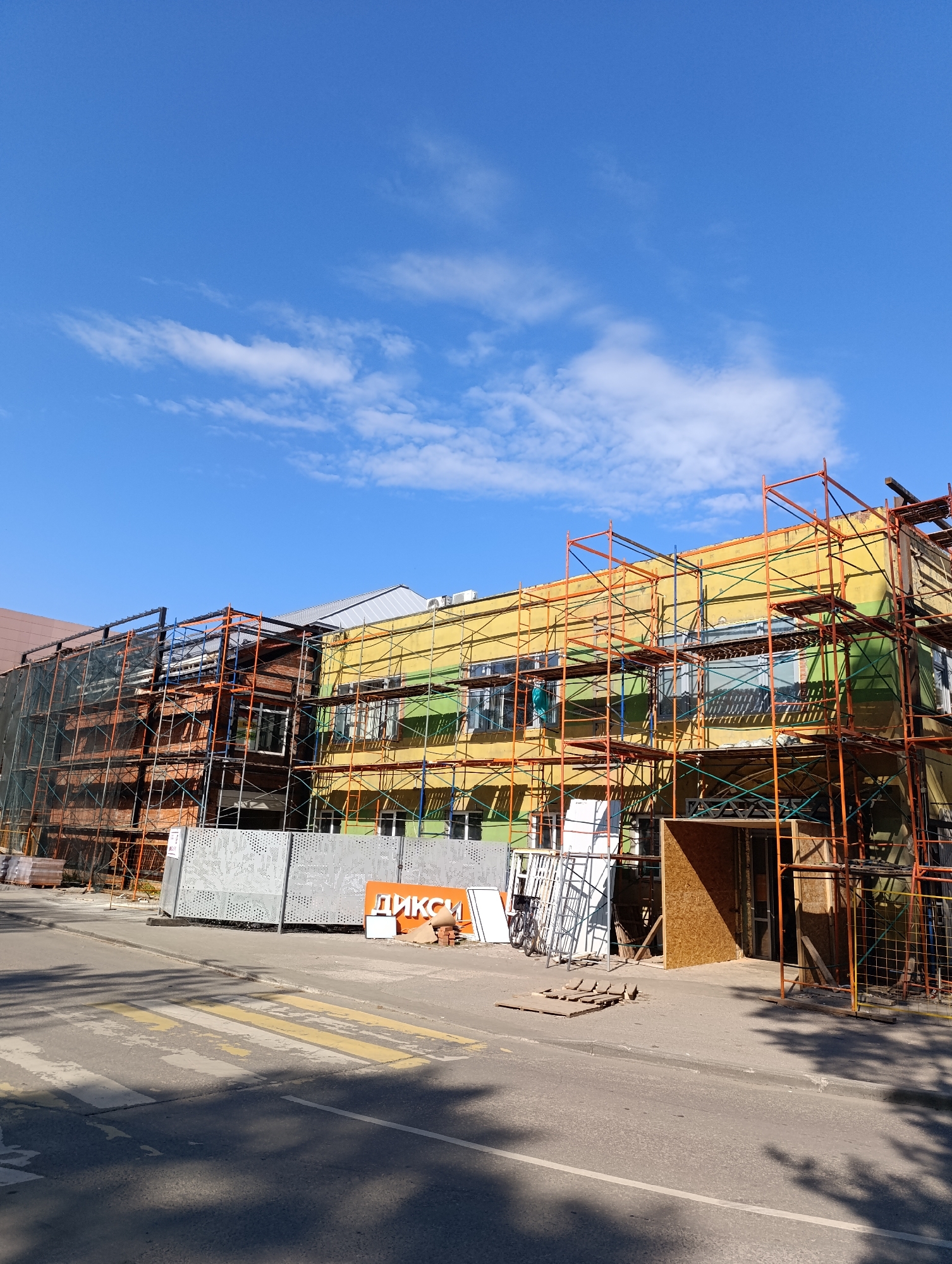 The image shows a three-story building in Unknown, Unknown, undergoing renovation. Scaffolding surrounds the exterior of the building, which is painted in sections of yellow, green, and brick. Construction materials and equipment are scattered around the base of the building. A sign with the word "ДИKCИ" is visible in front of the building. A crosswalk is painted on the street in the foreground. The sky is blue with scattered clouds.