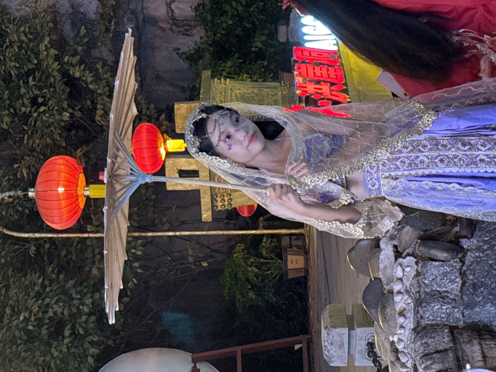 A young woman in a purple and gold costume with a veil stands holding a parasol. She has white face paint with purple accents. She is in an outdoor area with trees and traditional-style decor, including red lanterns and ornate structures. Behind her, there is a neon sign with Chinese characters. The setting appears to be part of a festival or cultural event in Yuzhong District, China.