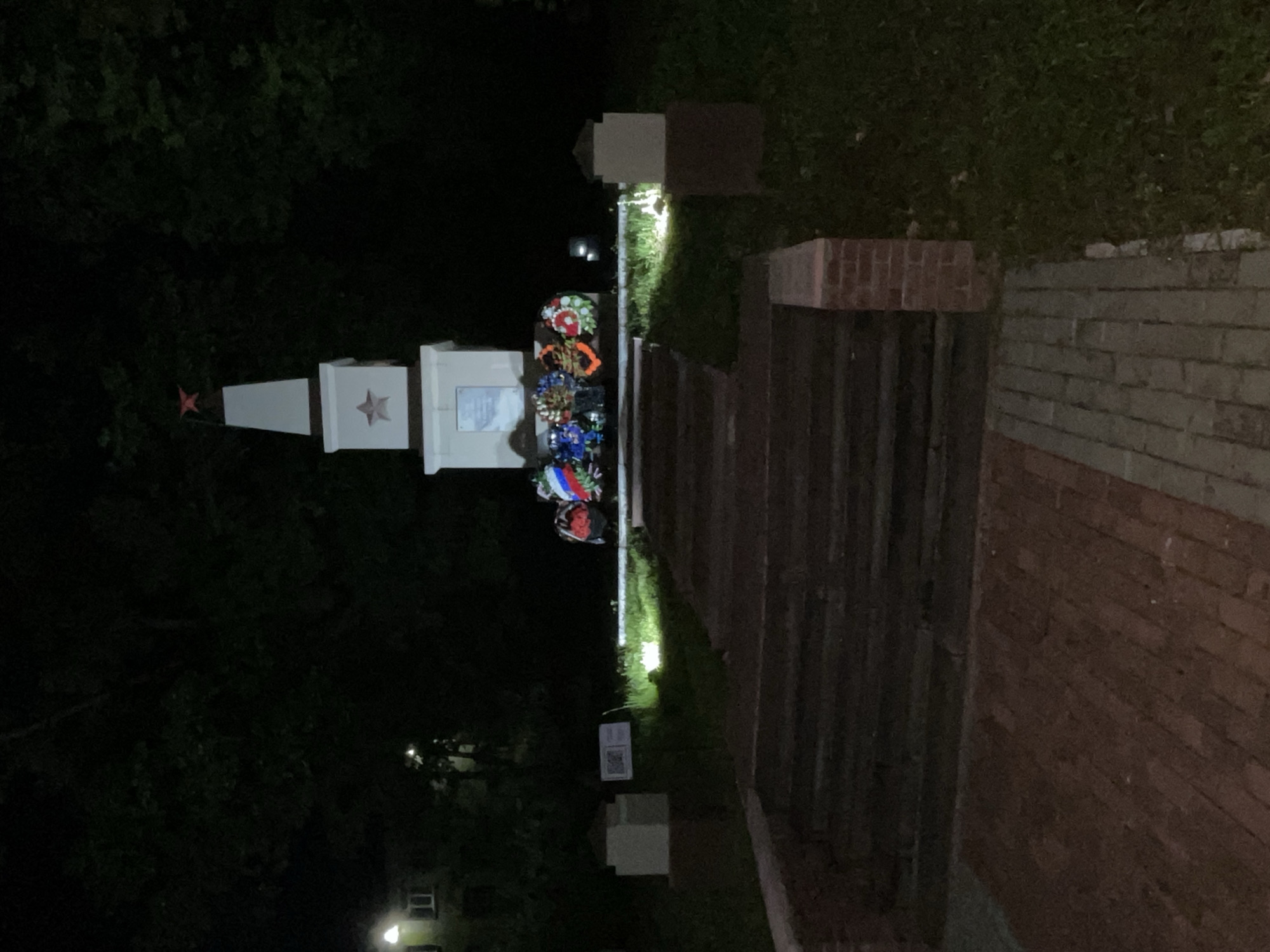 The image depicts a memorial or monument at night, located outdoors, likely in Красные Баки, Russia. The scene is dimly lit by artificial lights, highlighting specific elements against a dark background of trees and foliage.
The main subject is a multi-part, white-colored monument. On the left, a tall, slender obelisk-like structure tapers upwards, topped with a reddish-orange five-pointed star. To its right, a lower, rectangular block features a darker five-pointed star emblem. Adjacent to this block, another section holds what appears to be an inscribed plaque, though the text is illegible.
At the base of the monument, several large, circular wreaths made of artificial flowers are arranged. Some of these wreaths prominently display the colors of the Russian flag – white, blue, and red – indicating a national or patriotic commemoration.
To the right of the monument, a paved path, possibly constructed from reddish-brown bricks or pavers, extends towards the memorial. Ground-level lights illuminate sections of the path and the adjacent grassy areas. Further to the right, what appears to be a wooden fence or wall is visible, beyond which a brick wall can be discerned. The background consists of dark, dense trees and bushes. In the far distance, faint, glowing lights suggest the presence of other buildings or structures.
The time of day is clearly nighttime. No people are visible, and no specific activity is taking place other than the presentation of the wreaths. The overall atmosphere conveys solemnity and remembrance. A small white sign with an unreadable QR code and text is visible on the ground near the monument.