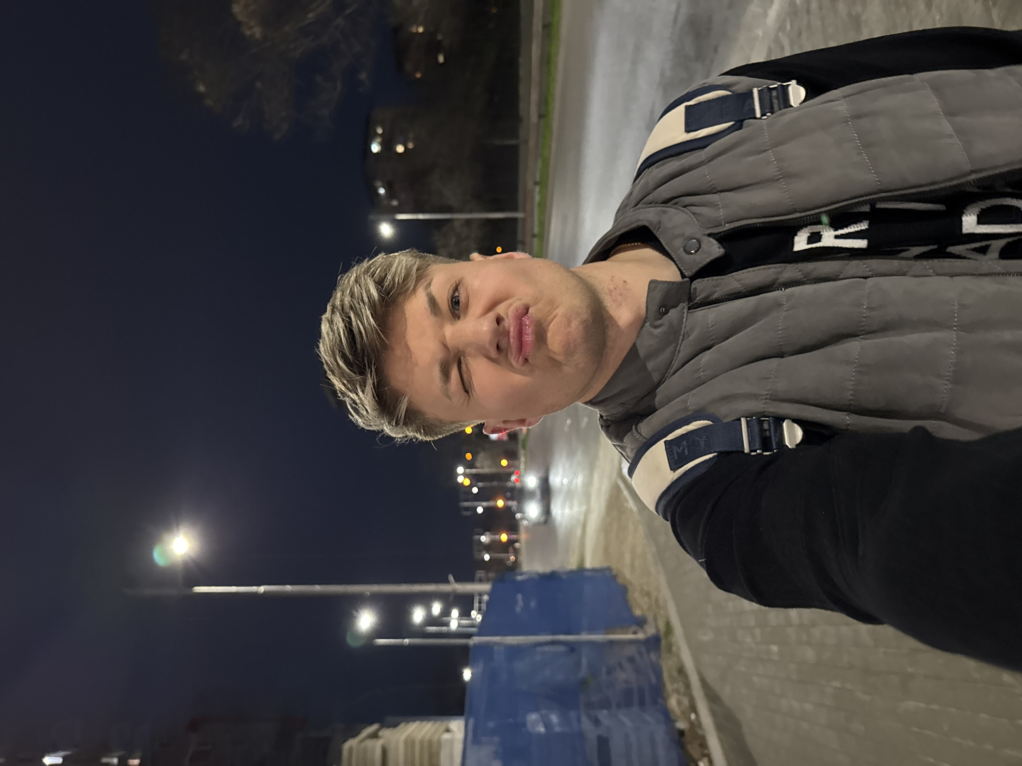 A person with blonde hair and a grey vest stands outdoors at night. The person is winking and pouting. Street lights illuminate the scene. The background shows a road with cars and buildings. There is blue construction netting. The location is likely a street in Plovdiv, Bulgaria.