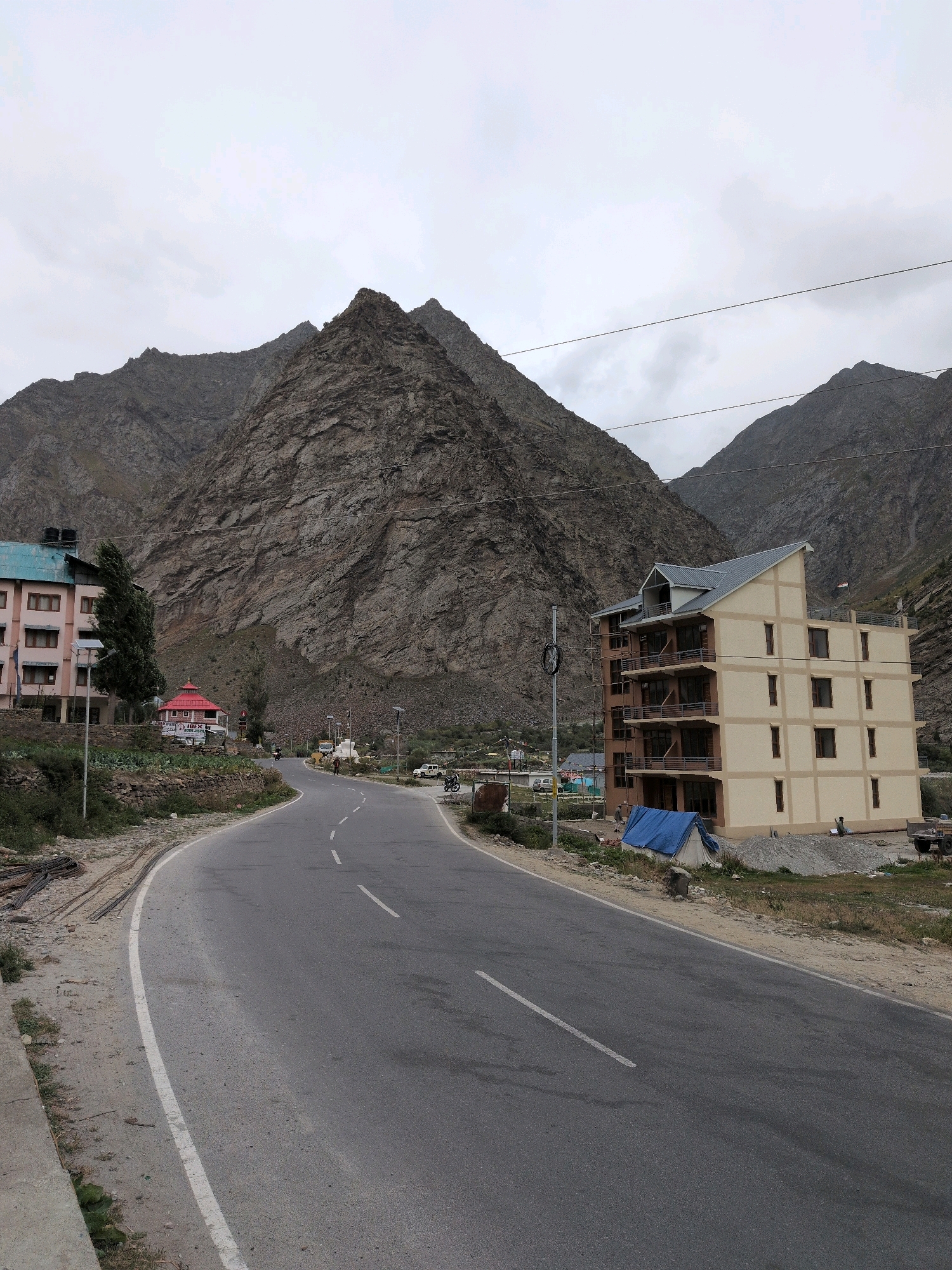 The image captures a scene in the city of Gemur, India. A winding asphalt road with white dashed lines dominates the foreground, curving from the left towards the right. To the left of the road, a light pink building with a turquoise roof and multiple windows is partially visible. Adjacent to this building, a tall, slender evergreen tree stands. Further down the road on the left, a small building with a bright red roof and traditional architecture is present.
On the right side of the road, a multi-story building with a beige facade and a gray, slanted roof is prominently featured. This building has multiple balconies and numerous windows. In front of this building, a blue tarp covers a tent-like structure, and a pile of gravel is visible. A person is observed near the building, and another person appears to be working on the roof.
The backdrop of the scene consists of towering, rugged mountains with visible geological striations, under a cloudy sky. Power lines are strung across the frame, running horizontally above the mountainous landscape. A few vehicles, including a motorcycle and a car, are parked along the roadside. The overall environment suggests a mountainous region with ongoing construction and development.