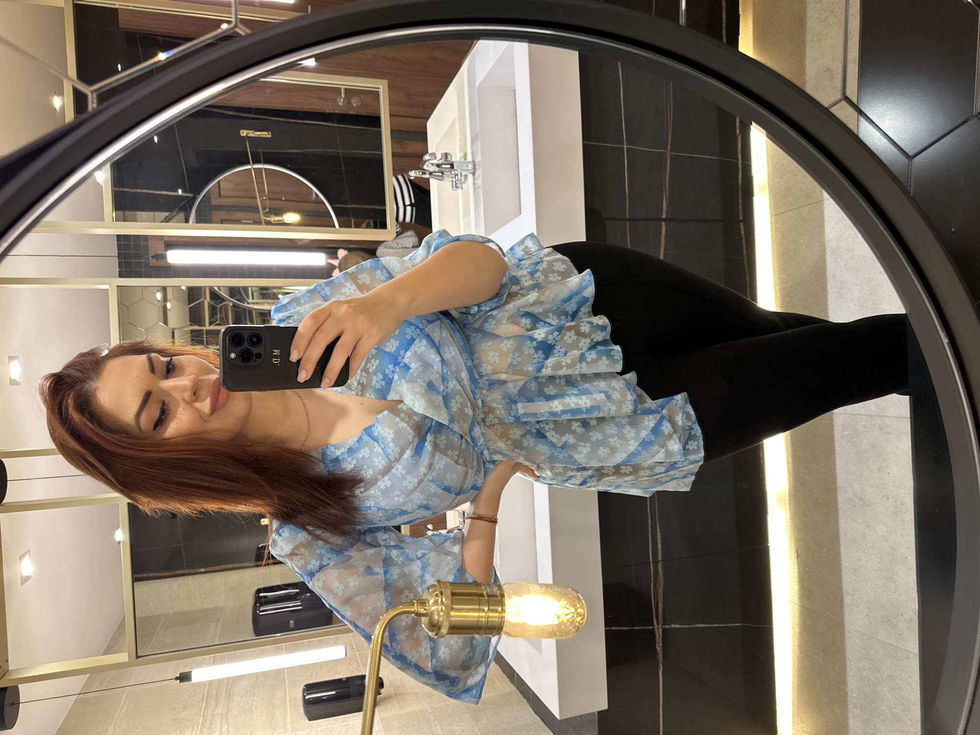 A young woman with auburn hair is taking a selfie in a bathroom mirror. She is wearing a blue floral-patterned top and black leggings. The bathroom features black and white tiles, a white sink, and a brass-colored lamp with a visible filament bulb. The image is taken in the city of Kazanlak, Bulgaria.