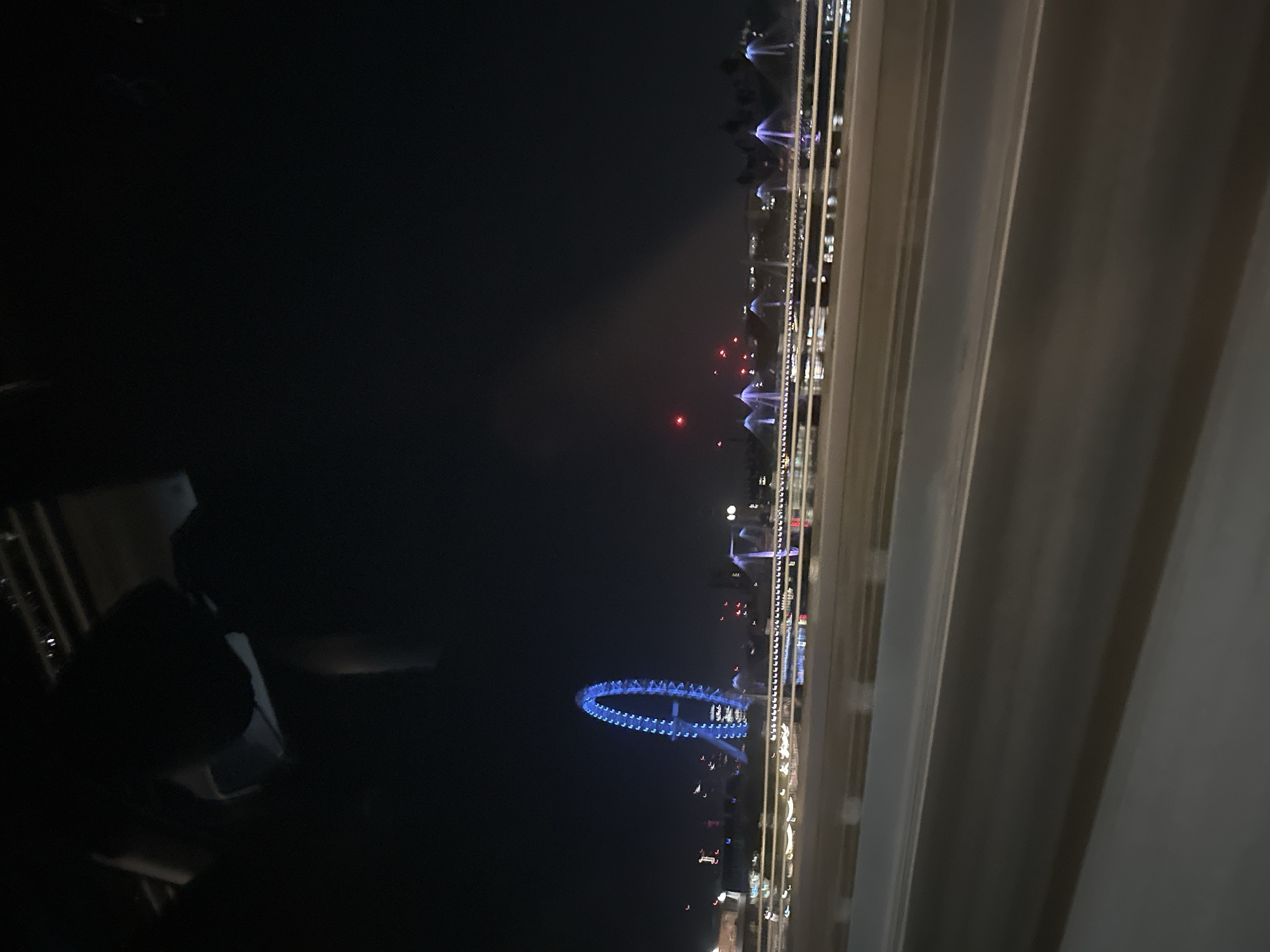 The image shows a night scene in London, United Kingdom, viewed from inside a building through a window. The London Eye is visible, illuminated in blue. A series of lights stretch along the river or a road, and some are arranged as fountains. The interior view includes parts of furniture, suggesting an observer sitting indoors.
