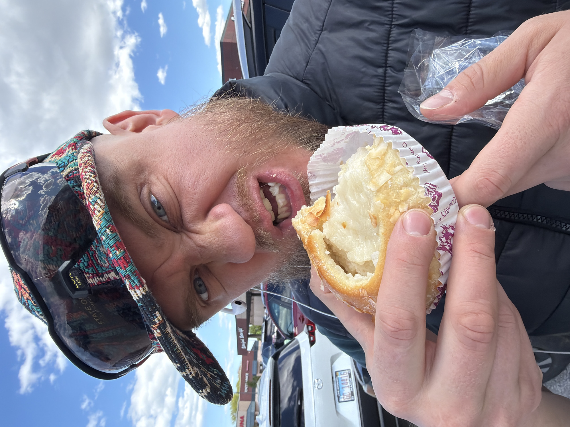A selfie shows a man with a beard wearing a multicolored cap and dark-colored puffy jacket. He is biting into a pastry inside a white cupcake wrapper. In the background, a white car with a license plate "GO 14416" is visible, along with other cars and buildings. The location is identifiable as the city of Niles, in the United States of America.