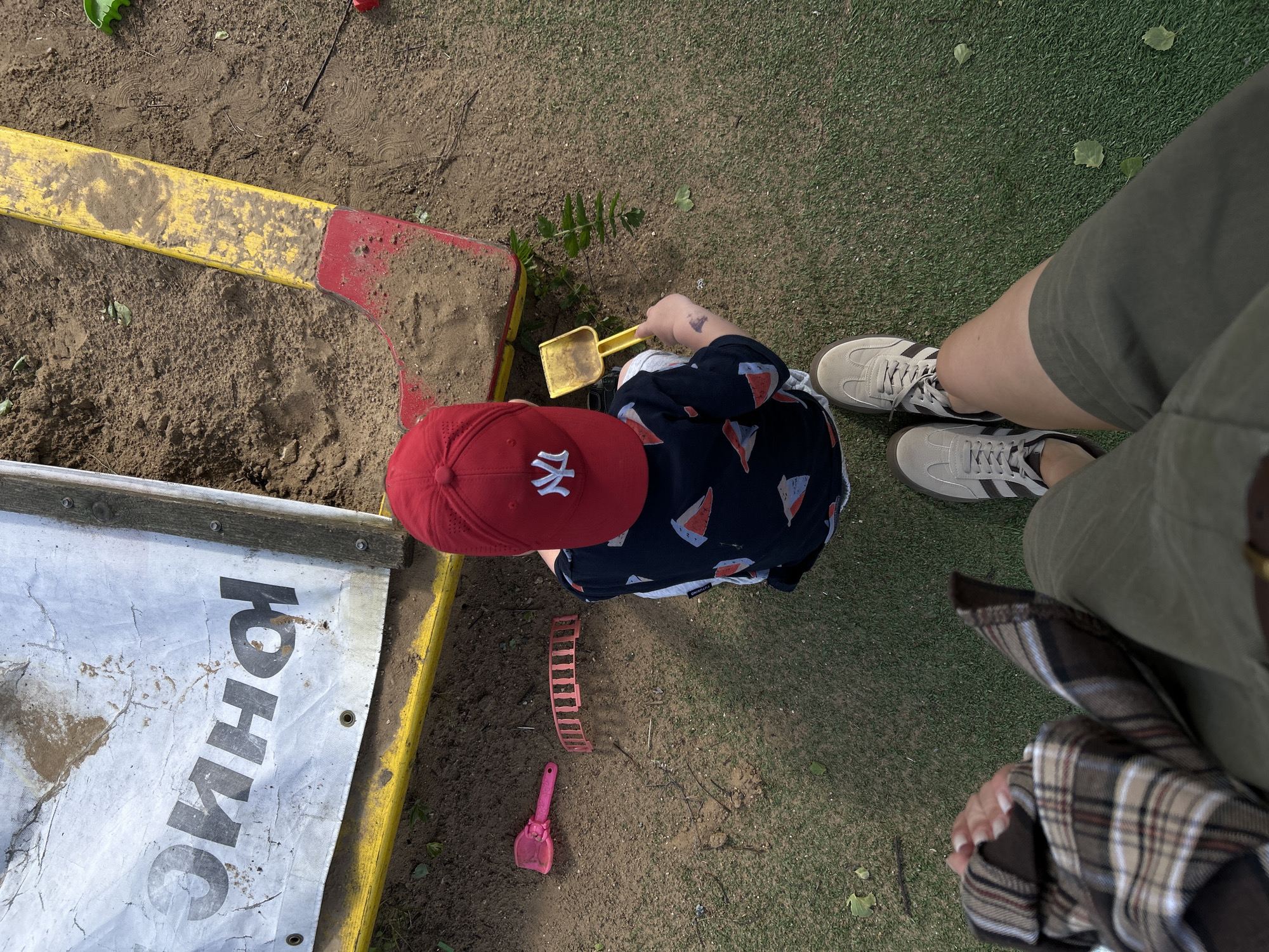 The image presents an overhead view of an outdoor scene. A child wearing a red baseball cap with a white logo, a dark shirt patterned with sailboats, and light-colored pants is playing in a sandpit. The child is holding a yellow plastic shovel. Around the sandpit are various toys, including a pink shovel and a red sand mold. The sandpit has a yellow and red rim on one side and a white and yellow rim on another side with Russian text on it. A portion of an adult's legs and shoes are visible on the right side of the image. The ground surrounding the sandpit is covered with artificial grass and some dirt. The location is outdoors, possibly in Борщевка Малая, Russia.