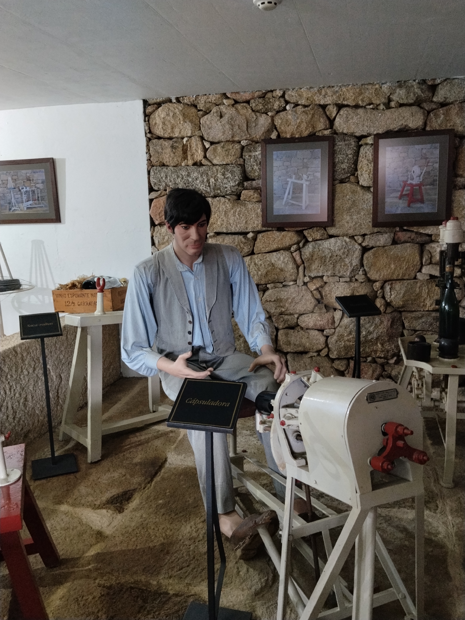 The image captures a static exhibit in Tarouca, Portugal, likely within a museum or cultural center. The scene is staged within a room that features a rough, uneven stone floor and a wall constructed from large, irregularly shaped stones. On the left side of the image, the wall transitions to a smooth, white surface.
The main subject of the image is a mannequin, dressed in a light blue button-down shirt, a grey vest, and light grey trousers. The mannequin is seated next to a piece of machinery, a white-painted device with red operational levers. A black sign with the word "Cápsuladora" in gold lettering is positioned next to the mannequin, indicating the machine's function - likely related to capsule application.
To the left, there's a wooden box filled with objects and labeled "VINHO ESPUMANTE NATURAL 12/2 GARRAFAS". Next to it, a black sign labeled "Saca-rolhas" stands on a metal pole.
Two framed pictures hang on the stone wall, depicting similar machinery or furniture pieces. A small white table with a green glass bottle and some other objects are seen in the background. The scene is indoors, and the lighting appears to be artificial.