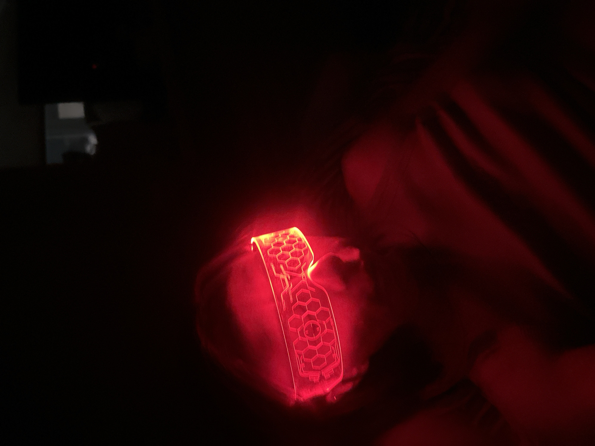 A close-up shot in a very dark room captures a person undergoing a red light therapy session. The central subject is a glowing red device, resembling a mask or facial applicator, worn by or held against the person's face. This device emits a vibrant red light, illuminating the immediate area around the person's head and casting a crimson glow on their skin and surrounding dark fabric, likely bedding or clothing.
The device itself is intricately designed with a prominent honeycomb or hexagonal pattern. Within these geometric shapes, etched lines resembling circuitry are visible. Notable symbols on the device include a stylized 'R' or similar emblem and a cross or plus sign. Parts of the person's hand are visible near the device, suggesting they are either holding it or adjusting its position.
The setting is an indoor environment, almost entirely dark except for the light from the therapy device, indicating it's likely night or a room with lights off. The deep shadows obscure most background details, with only faint, indistinct shapes barely visible in the top-left background. There are no specific cues to identify the location as Votkinsk, Russia, within the frame. The image conveys a private and therapeutic activity.
