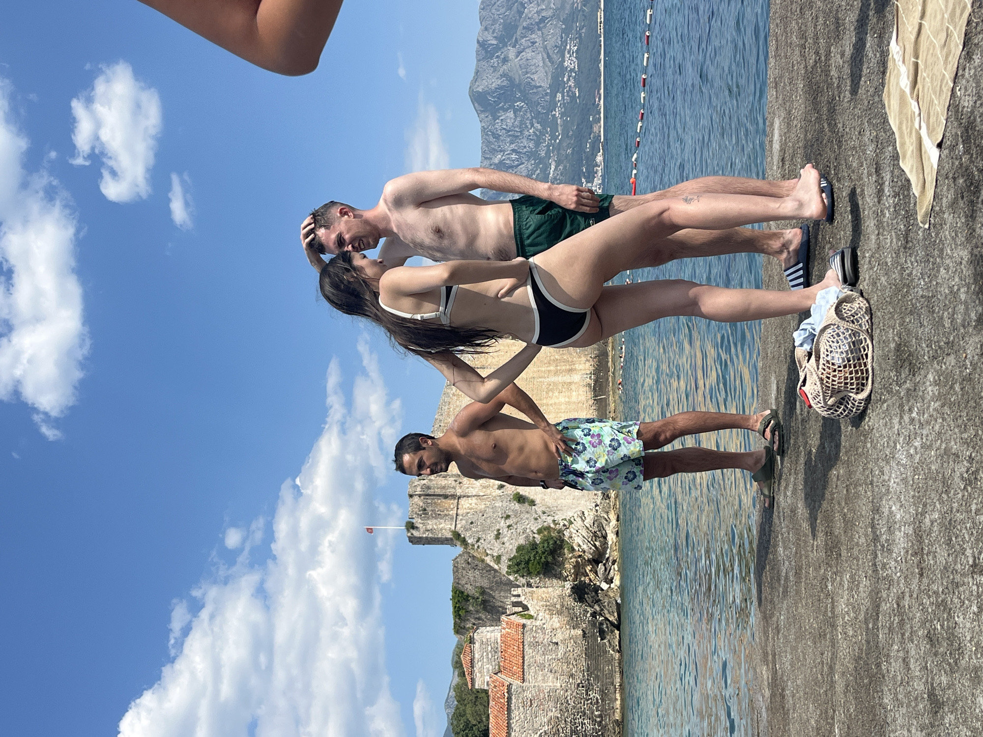 This media file captures a sunny day by the sea in Budva, Montenegro. Three people are the main subjects, standing on a concrete pier overlooking the clear blue water. The image is oriented vertically, but the scene itself suggests a horizontal composition.
In the foreground, a man and a woman are depicted in an intimate embrace. The woman wears a black and white bikini, and the man is shirtless, wearing dark green swim trunks. They appear to be close, with the man touching his own head and the woman's hand on his chest. Their focus is on each other, suggesting a moment of affection.
Behind them, a second man is positioned lower in the frame, also shirtless and wearing colorful floral swim trunks. He is looking towards the left of the frame and appears to be standing further away from the couple.
In the background, a historic stone fortress, likely a landmark in Budva, is visible on the left. The fortress features terracotta-tiled roofs and ancient stone walls, with a flag flying on a pole. The sea stretches out to the right, with a rope barrier marking a swimming area. The sky is a bright blue with fluffy white clouds, indicating a clear and pleasant day. The sun is high in the sky, suggesting it is midday. The overall atmosphere is relaxed and summery. There is no visible text in the image.
