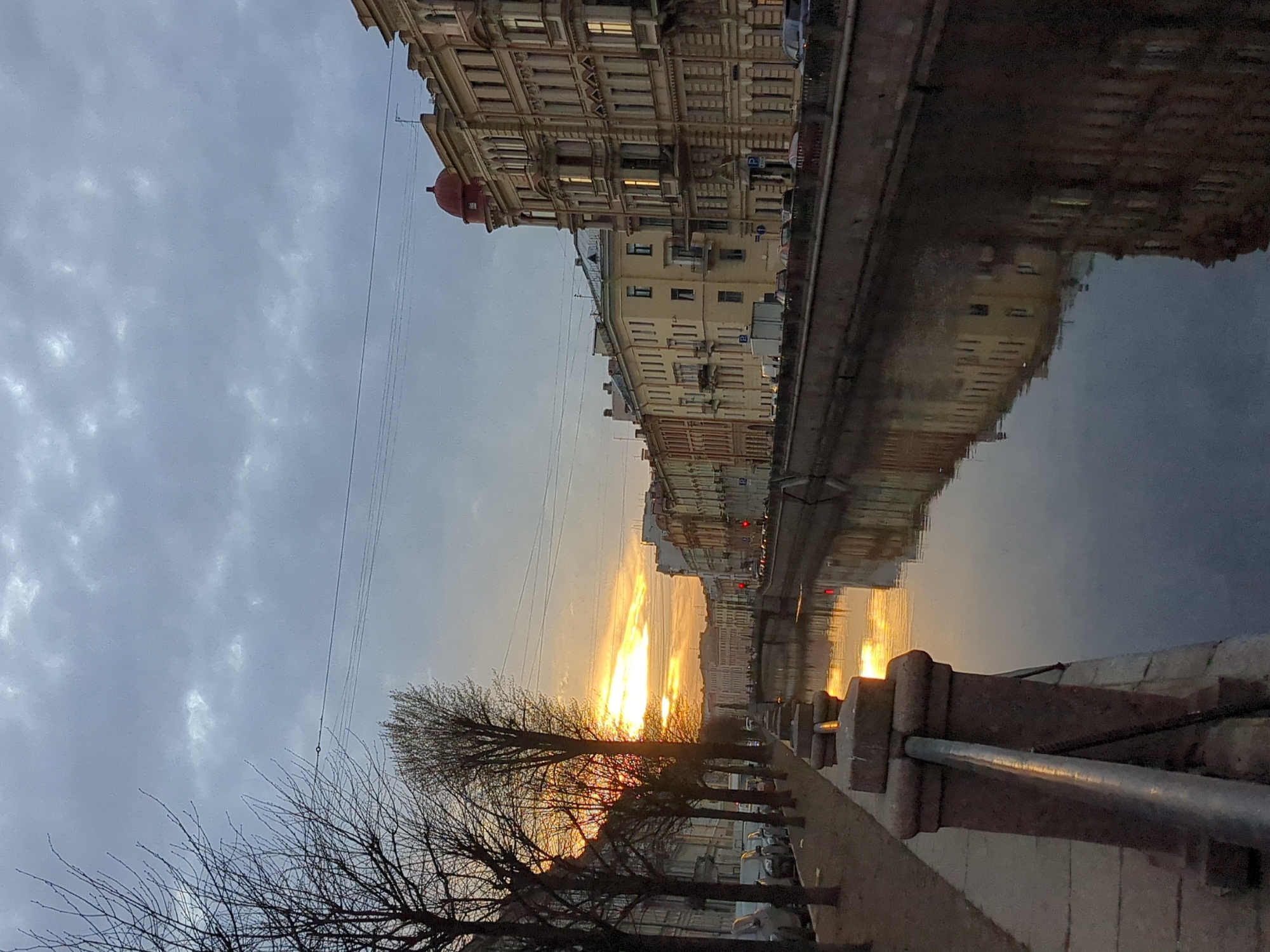 The image shows a canal in Saint Petersburg, Russia, at sunset. The sky is filled with gray clouds, but the setting sun casts a golden glow on the buildings lining the canal. The buildings are reflected in the water. Bare trees line the walkway along the canal. A metal railing runs along the edge of the walkway.