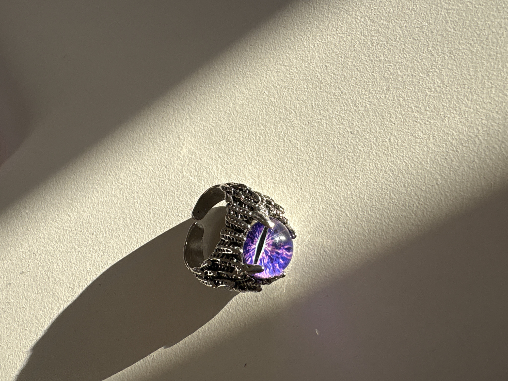 The image shows a close-up, eye-level shot of a silver ring with a purple, iridescent glass gem designed to look like an eye. The ring band is intricately detailed with a dark, metallic texture that appears to resemble claws or tendrils wrapping around the gem. The ring is resting on a textured, off-white surface. A strong light source from the left casts a long, sharp shadow diagonally across the surface and the ring. The lighting creates a high-contrast scene, with the ring and its shadow being the primary focal points. There is no indication of the city of Kaliningrad, Russia, in the image.