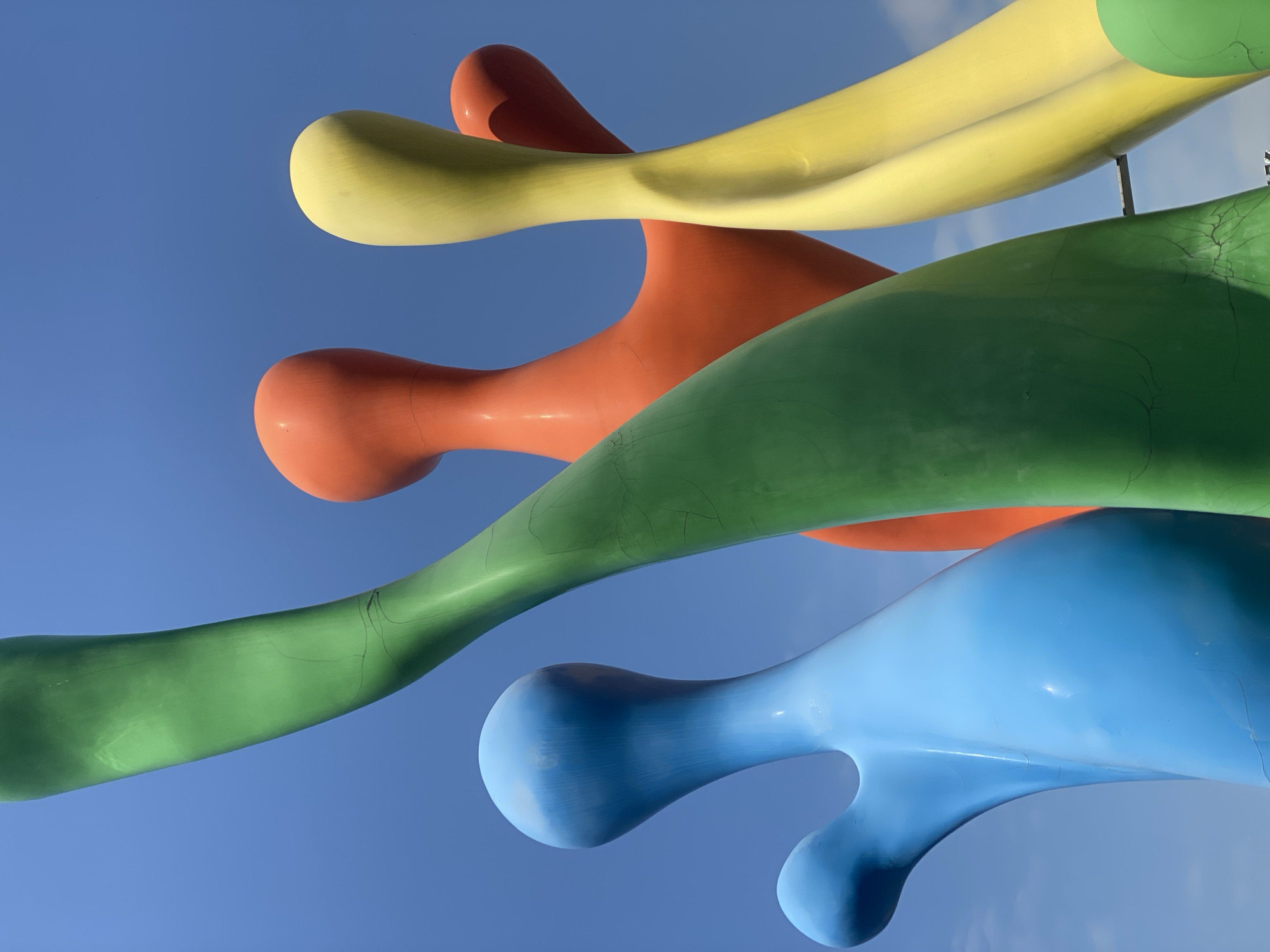 This is a low-angle, medium shot of a large, colorful outdoor sculpture against a bright blue sky. The sculpture appears to be made of smooth, polished material and consists of abstract, bulbous forms in vibrant shades of blue, green, orange, and yellow. The blue form is on the left, the green form dominates the center, and orange and yellow forms are visible on the right, with a smaller orange form peeking out from behind the yellow. Some minor cracks are visible on the surface of the green and orange elements. The overall impression is one of abstract, playful shapes reaching towards the sky. The specific location in Novokuznetsk, Russia is not discernible from the image itself.