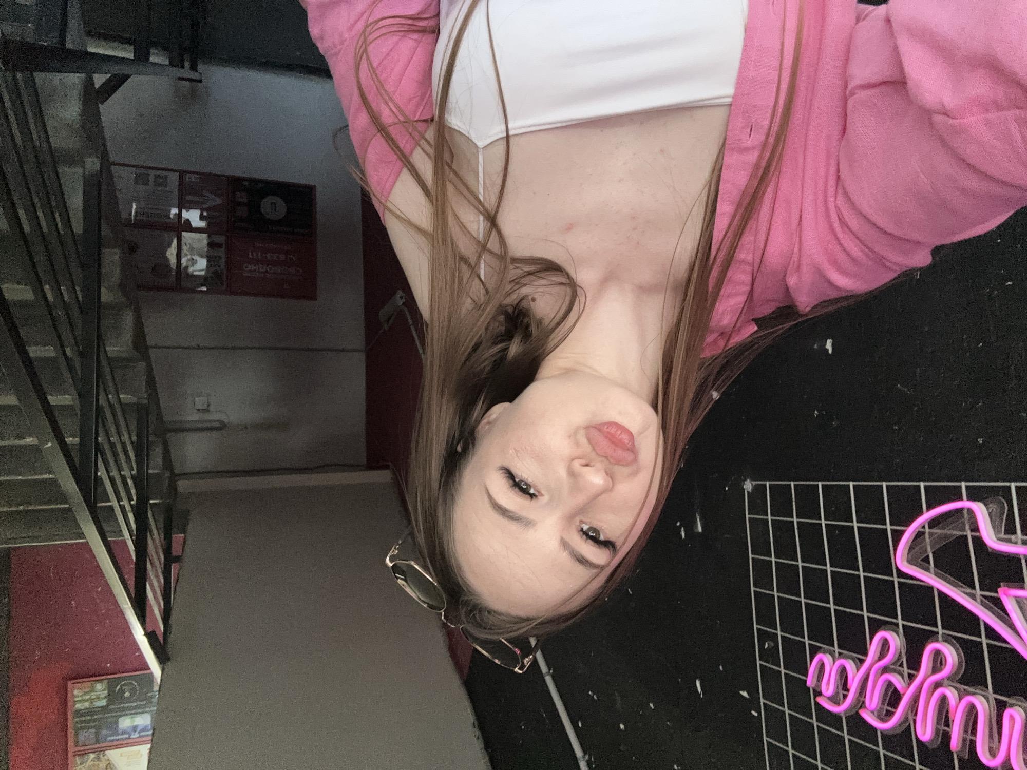 A young woman is depicted in a close-up, selfie-style photograph, looking directly at the camera with a slightly pouting expression. She has long, straight brown hair cascading over her shoulders and chest. She wears a white spaghetti-strap crop top layered under an open, bright pink cardigan or jacket. A pair of gold-rimmed glasses hangs from her neck, resting against her chest.
The setting appears to be an indoor space, possibly a commercial establishment like a bar or entertainment venue. To the left of the woman, a dark metal staircase railing is visible, ascending alongside a wall that is light-colored at the top and transitions to a deep red lower down. Further into the background on the left wall, several signs or posters are affixed; one prominently features an arrow pointing to the right, and another red sign contains text that is largely unreadable, though some numbers or letters might be present.
To the woman's right, a black wall with a distinct white grid pattern is featured. Against this, a pink neon sign is partially visible, appearing to spell out words in a stylized cursive font. The overall lighting in the scene is somewhat dim, suggesting an indoor evening or low-light environment. The woman is the primary subject, posing for the camera.