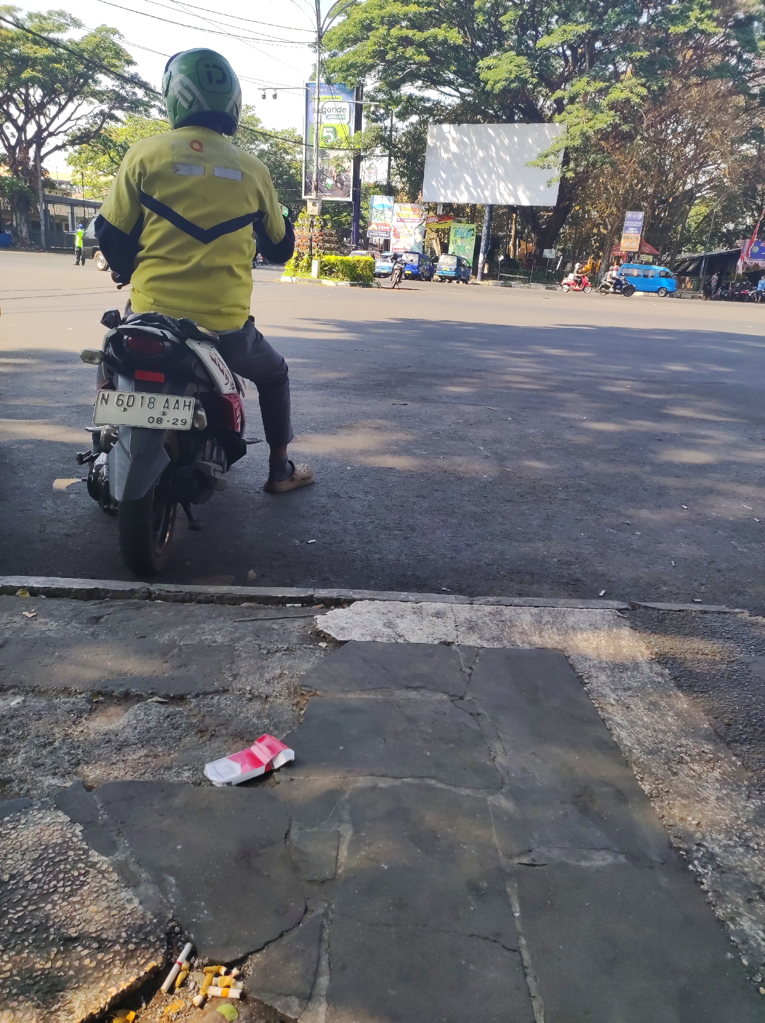 An individual wearing a green helmet and a yellow jacket with a dark blue V-shaped stripe is seated on a motorcycle, facing away from the camera. The motorcycle has a license plate that reads "N 6018 AAH 08-29". In the foreground, on a cracked pavement, lies a discarded red and white cigarette pack and several cigarette butts. The scene appears to be a street in Malang, Indonesia, indicated by the license plate format and common street signs visible in the background. Trees line the street, and other vehicles, including a blue van, are present further down the road. The lighting suggests it is daytime, with shadows cast across the pavement.