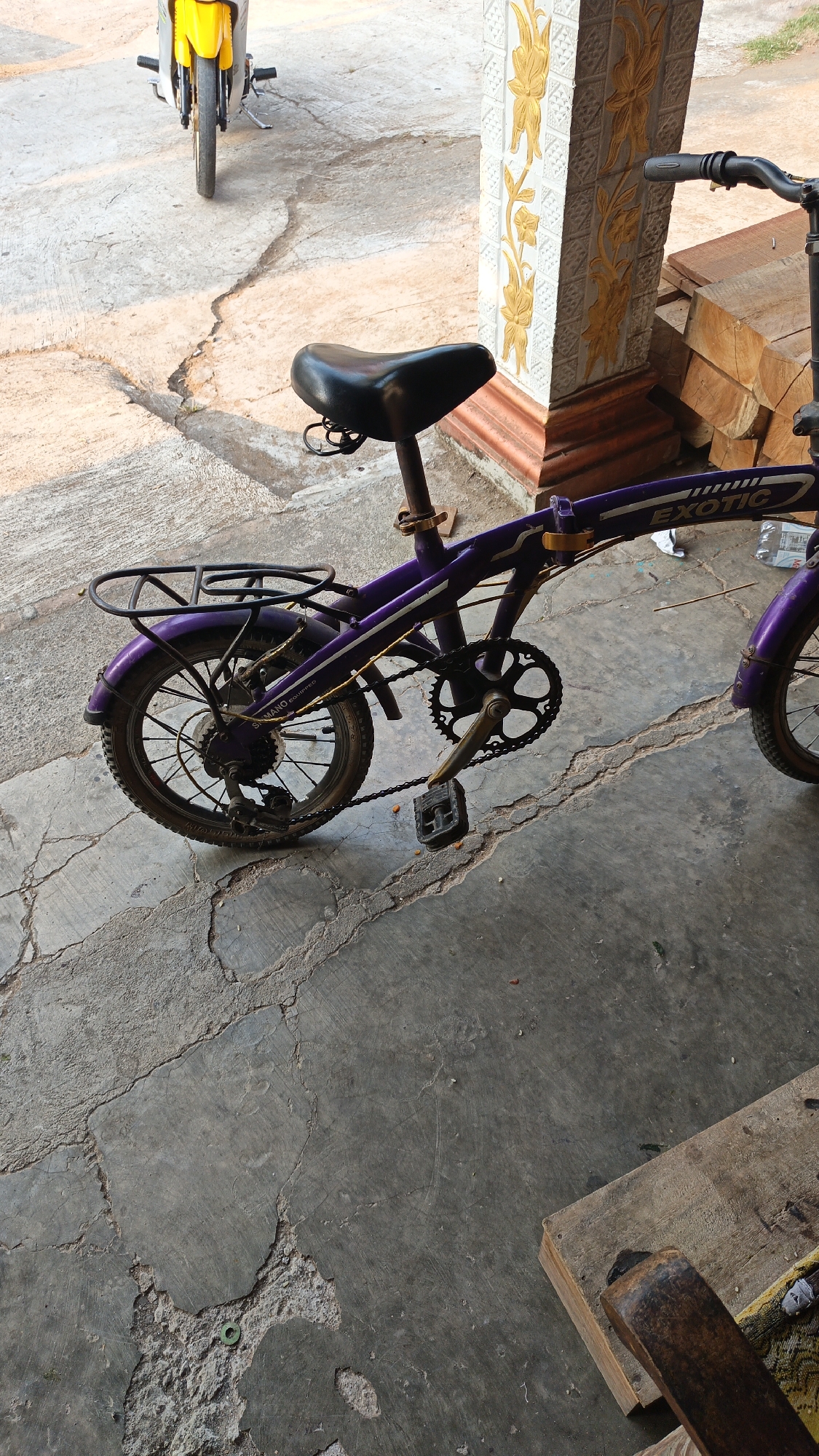 A purple folding bicycle with a black seat is parked on a cracked concrete surface. The bicycle has a rear rack and a chain with visible teeth on the crankset. To the left of the bicycle, a portion of a yellow and silver motorcycle is visible, parked on the same concrete surface. In the background, a decorative pillar with raised gold floral patterns stands next to a stack of cut lumber. The scene appears to be outdoors, possibly in a residential or workshop area in Sambi, Indonesia, given the presence of common Indonesian building materials and the overall aesthetic. The lighting suggests it is daytime, with warm sunlight casting shadows. There are no people visible in the image.
