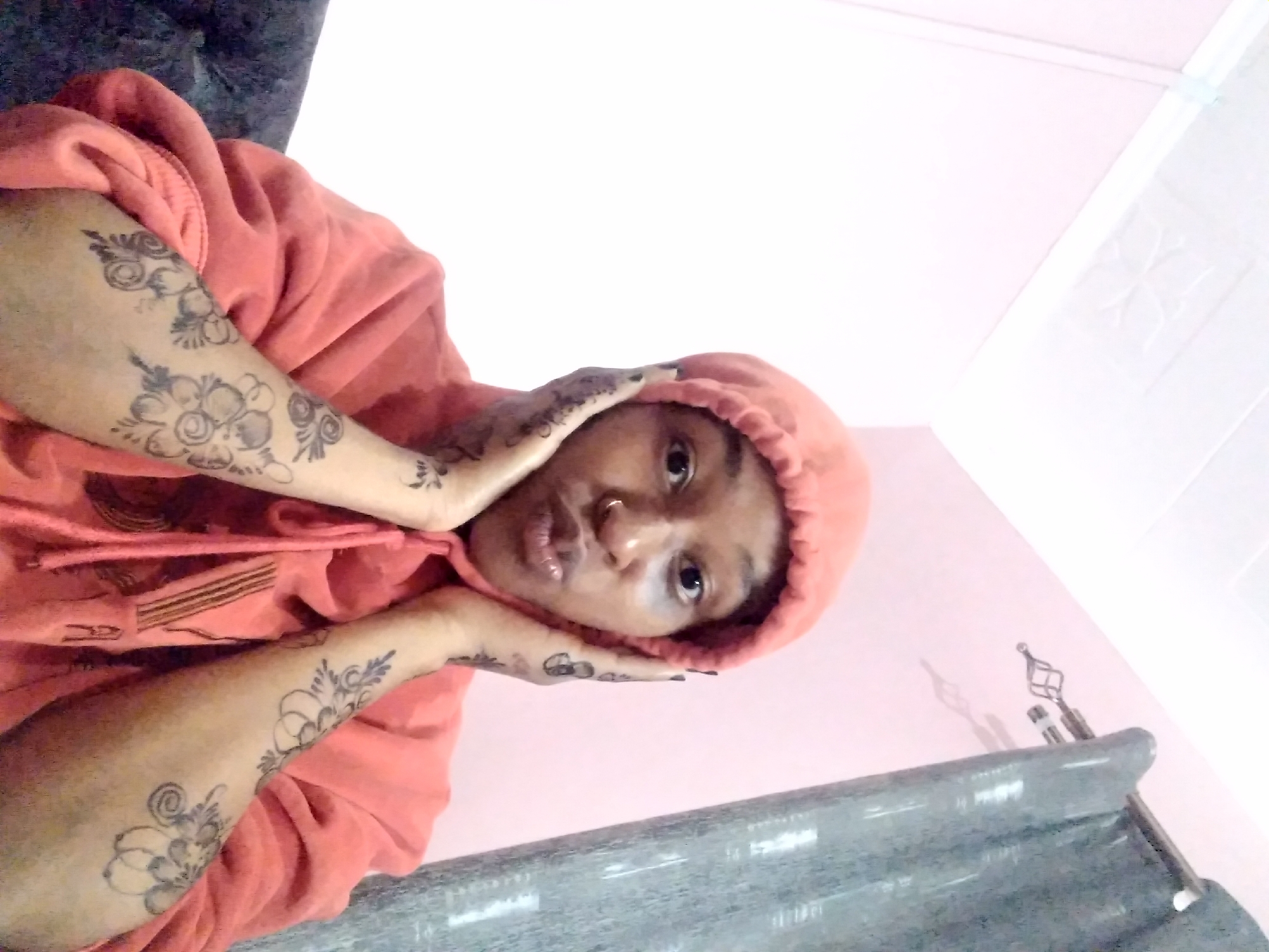 A close-up, canted shot features a person wearing a salmon-colored hooded sweatshirt, holding their face with both hands. The person has dark hair under the hood and a nose ring. Floral tattoos are visible on both forearms. The background consists of a pink wall and a white ceiling. A gray, textured surface is present at the bottom of the frame, and a curtain rod is visible in the bottom right corner. The location is Unknown, Unknown.