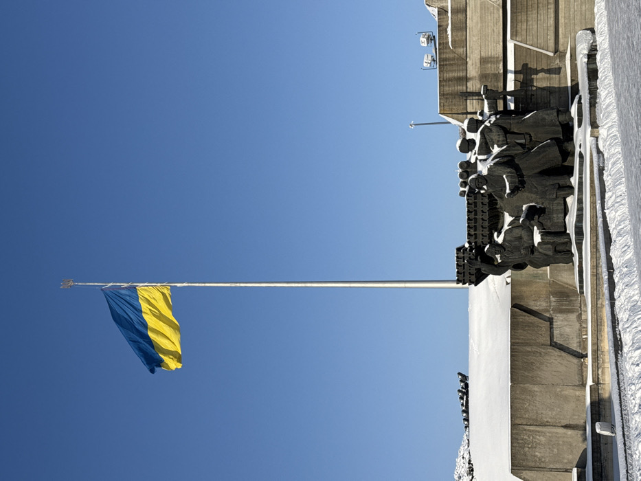 The image captures a clear winter day at the National Museum of the History of Ukraine in the Second World War complex, located on the Pechersk Hills in Kyiv, Ukraine. A very tall, slender silver flagpole, topped with a golden Tryzub (Ukrainian trident) finial, stands prominently in the upper left quadrant. From this flagpole, a large Ukrainian national flag, with its upper blue and lower yellow horizontal bands, is fully unfurled and ripples to the left against an uninterrupted, brilliant azure sky.
In the lower third of the image, a dark, multi-figure bronze sculptural group is visible. This group consists of approximately six figures, depicted in what appear to be military or worker uniforms, with one figure raising an arm. Behind and slightly above these figures, a horizontal arrangement of dark, cylindrical structures, resembling rocket launchers, is integrated into the monument. The sculpture is situated on a multi-tiered concrete base, which features angular, geometric patterns. The ground surrounding the monument, as well as the horizontal surfaces of the concrete structures, are uniformly covered in a layer of white snow. In the immediate foreground, a snowy pathway or cleared area is visible. To the right, additional concrete architectural elements, some with mounted equipment (possibly cameras or lights), are partially covered in snow.