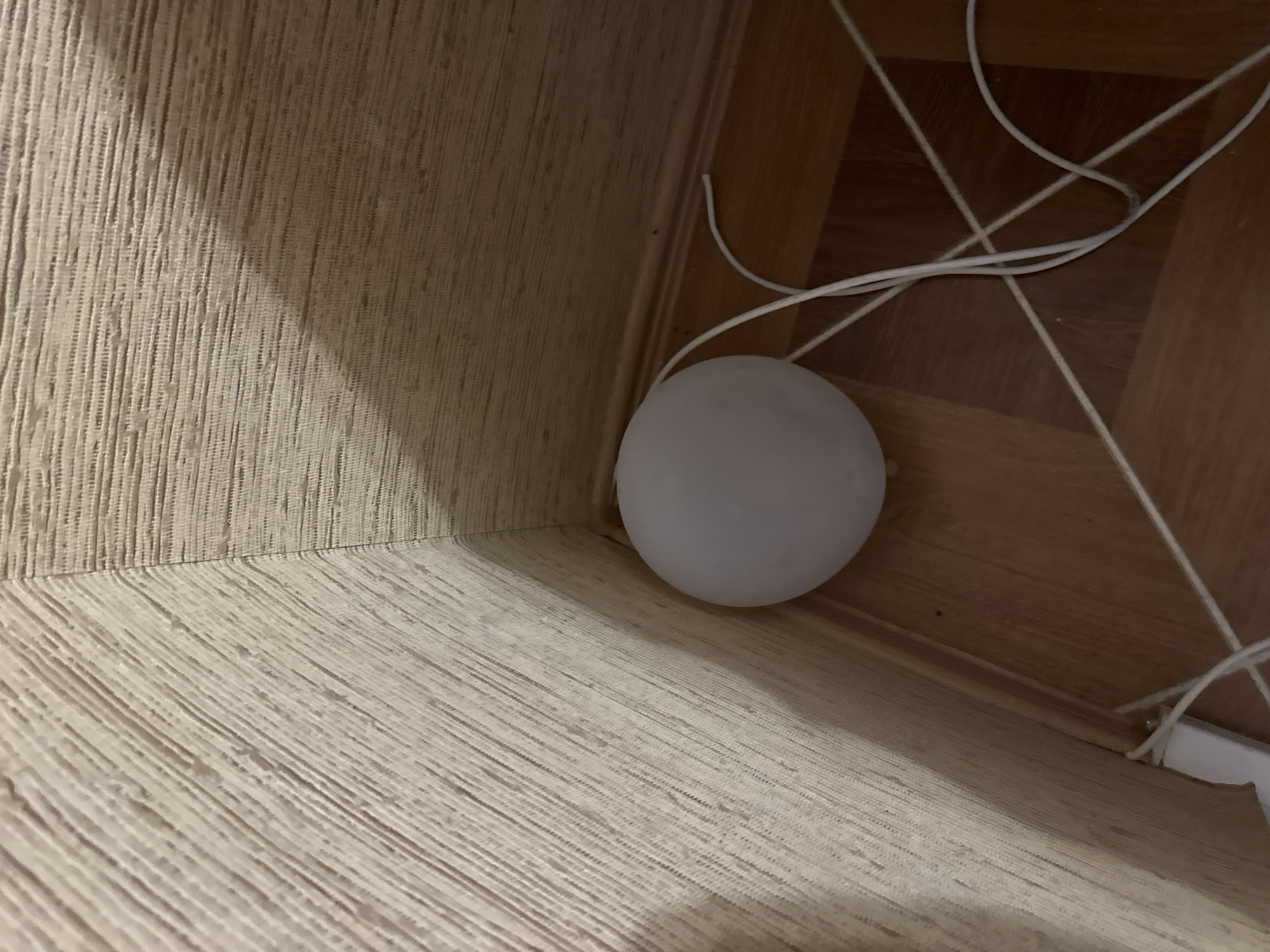 The image depicts a white, spherical object, possibly a light, placed in the corner of a room. The walls are covered with a textured, light-colored wallpaper. The floor is made of wooden planks. White electrical cords are visible on the floor, connected to the object. The scene is indoors and appears to be a residential setting. No individuals are present in the image. The lighting suggests it is either nighttime or the room is dimly lit. There is no specific indication of the city of Aleksandrovka, Russia, within the image itself.