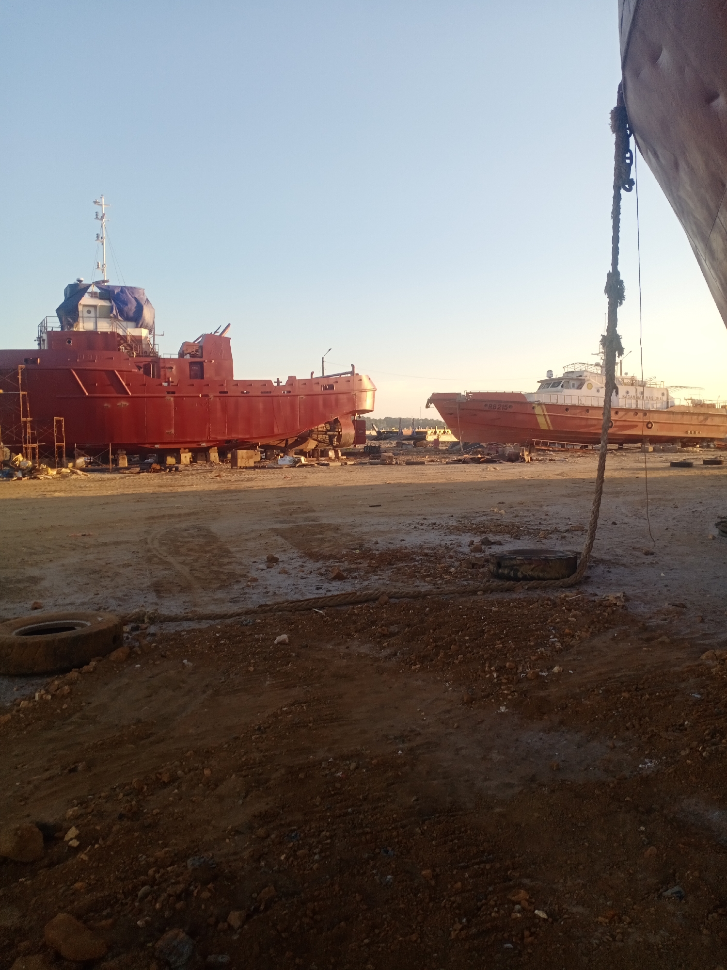 In Siddo, Indonesia, an industrial scene unfolds at a shipyard under a clear sky. Two large, red ships are the central focus. The ship on the left is incomplete, with scaffolding attached and a blue tarp covering its upper section. It rests on blocks, indicating it's undergoing construction or repair. To its right, a second, orange and white ship is more complete, with "RB 215" visible on its hull, along with the text "BADAN NASIONAL" on its side. This ship also appears to be out of the water, supported by a similar block system. The ground is a mixture of dirt and gravel, with scattered debris including tires and ropes. A thick, weathered rope hangs from the right foreground, suggesting active work or the presence of mooring equipment. The overall impression is one of maritime industry activity, with ships in various stages of readiness.