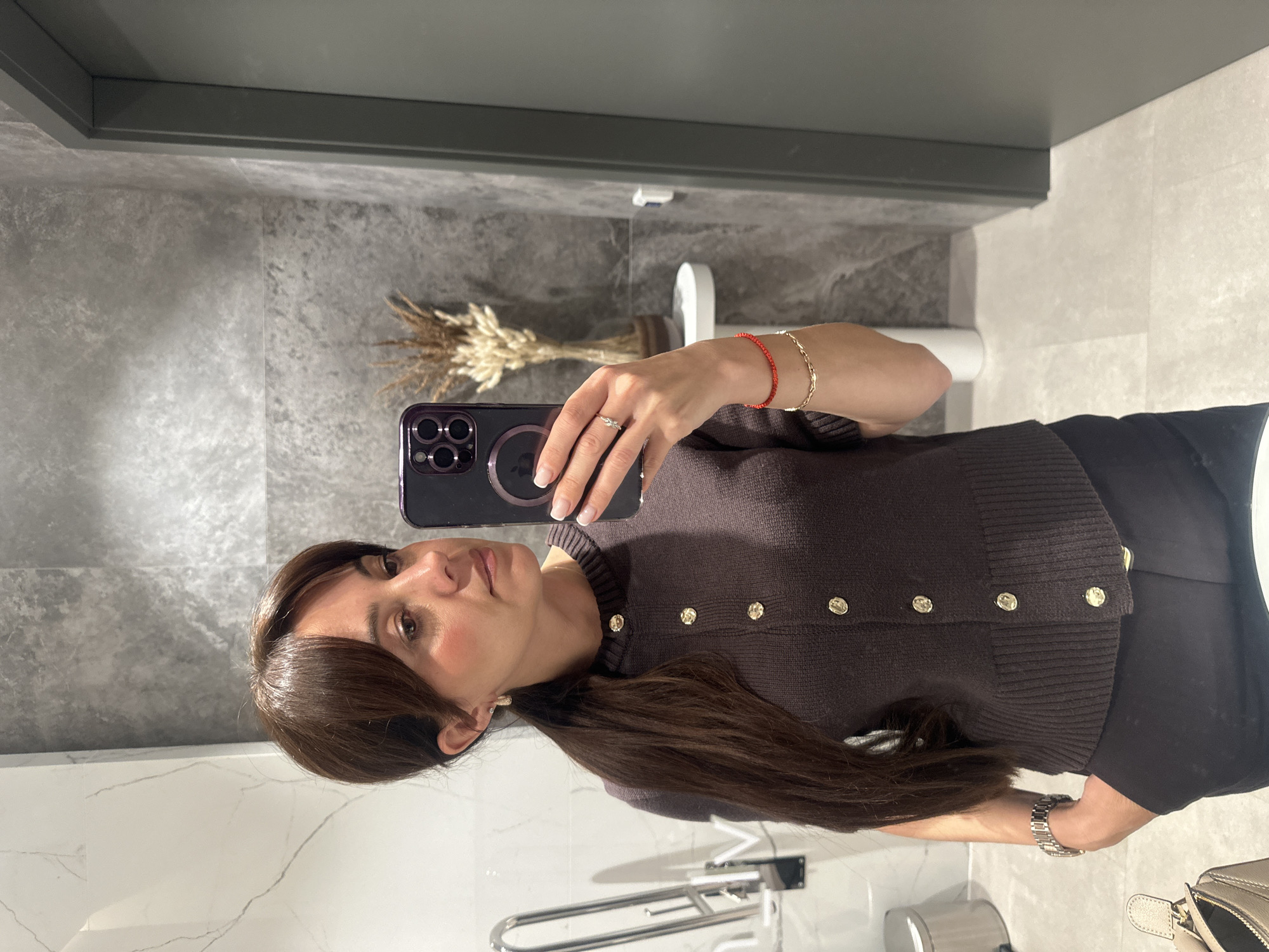 A woman is taking a selfie in a bathroom. She is holding a dark smartphone with multiple camera lenses and a MagSafe ring. The woman has shoulder-length brown hair with bangs, a brown cardigan with gold buttons, and is wearing bracelets and a ring. The bathroom has gray and white marbled walls and a gray ceiling. In the background, there is a bouquet of dried flowers in a brown vase, a white toilet, and grab bars for accessibility. The scene suggests a modern, well-maintained bathroom, likely in a public space or a commercial establishment. There is no direct evidence suggesting the location is Riga, Latvia.