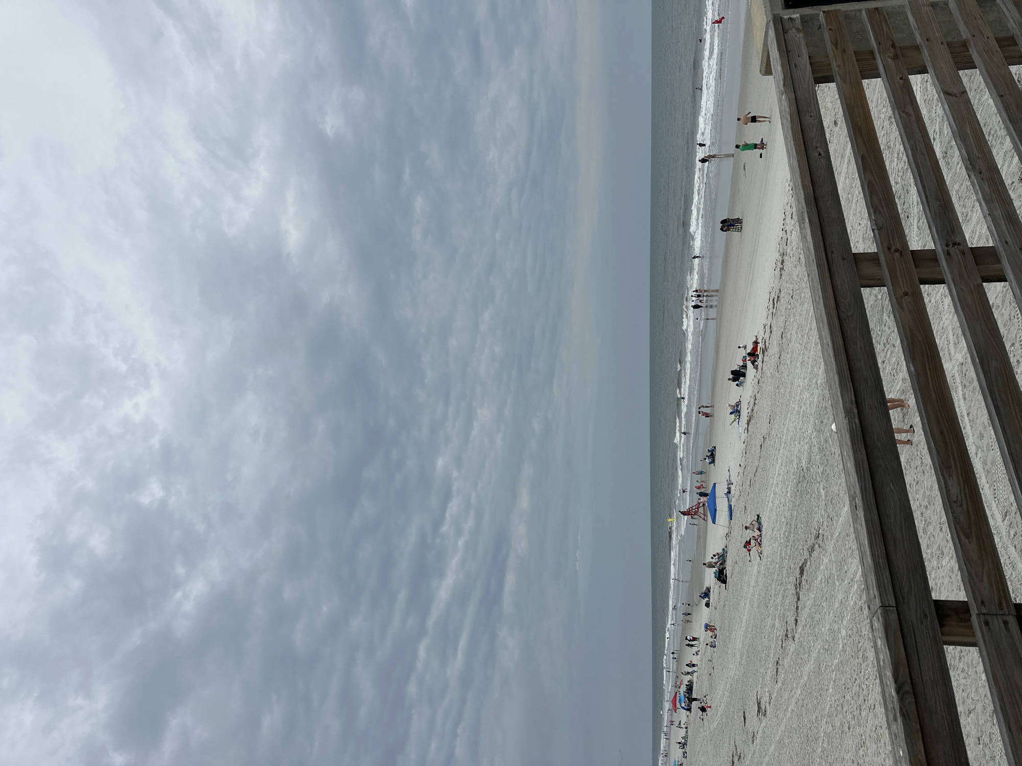 The image shows a beach scene in Jacksonville Beach, United States of America, taken from an elevated perspective near a wooden fence or walkway. The beach is crowded with people engaged in various activities like swimming, sunbathing, and walking along the shore. The sky is overcast with clouds. The horizon line is slightly blurry, possibly due to atmospheric conditions. The wooden fence runs along the right edge of the image, partly obscuring the view of the beach.
